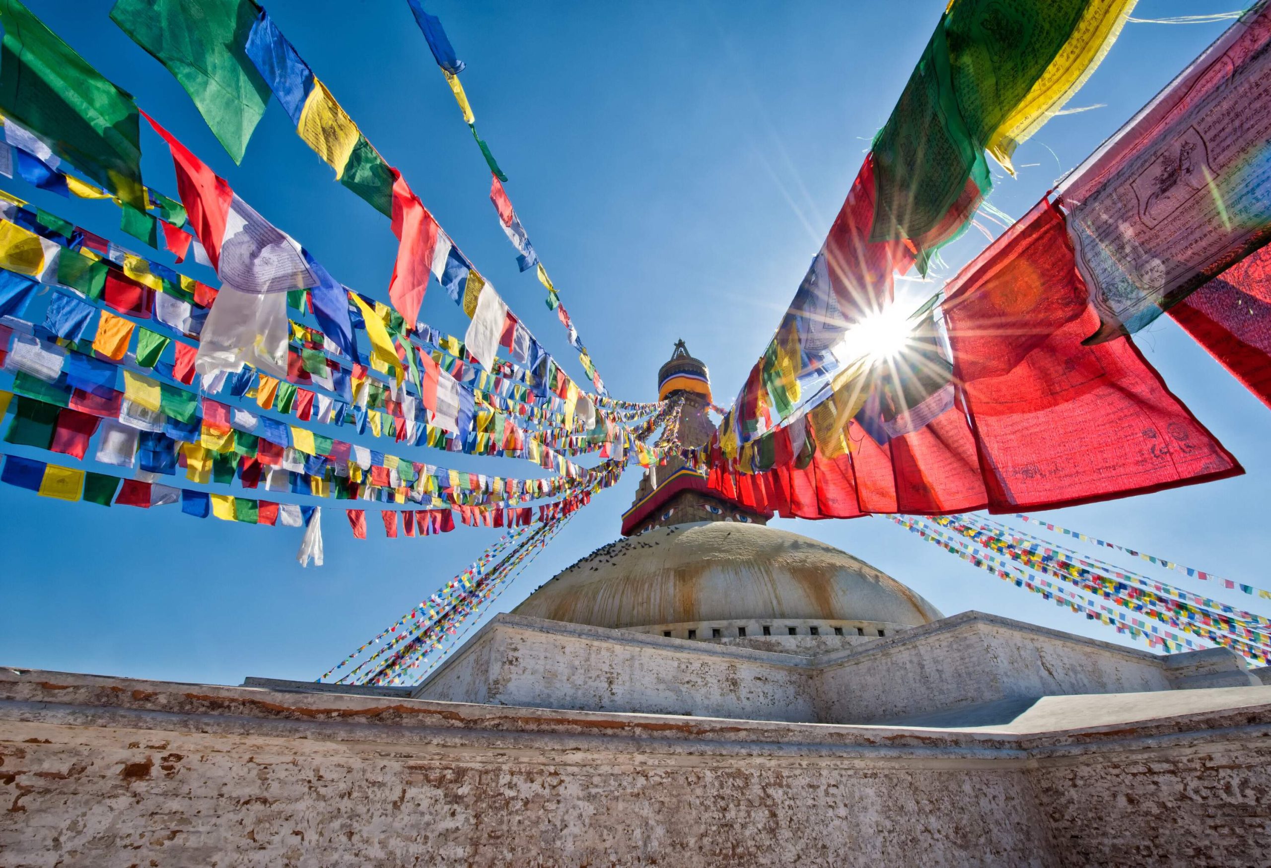 On top of a stupa are hanging colourful prayer flags against the sun's rays in the cloudless sky.