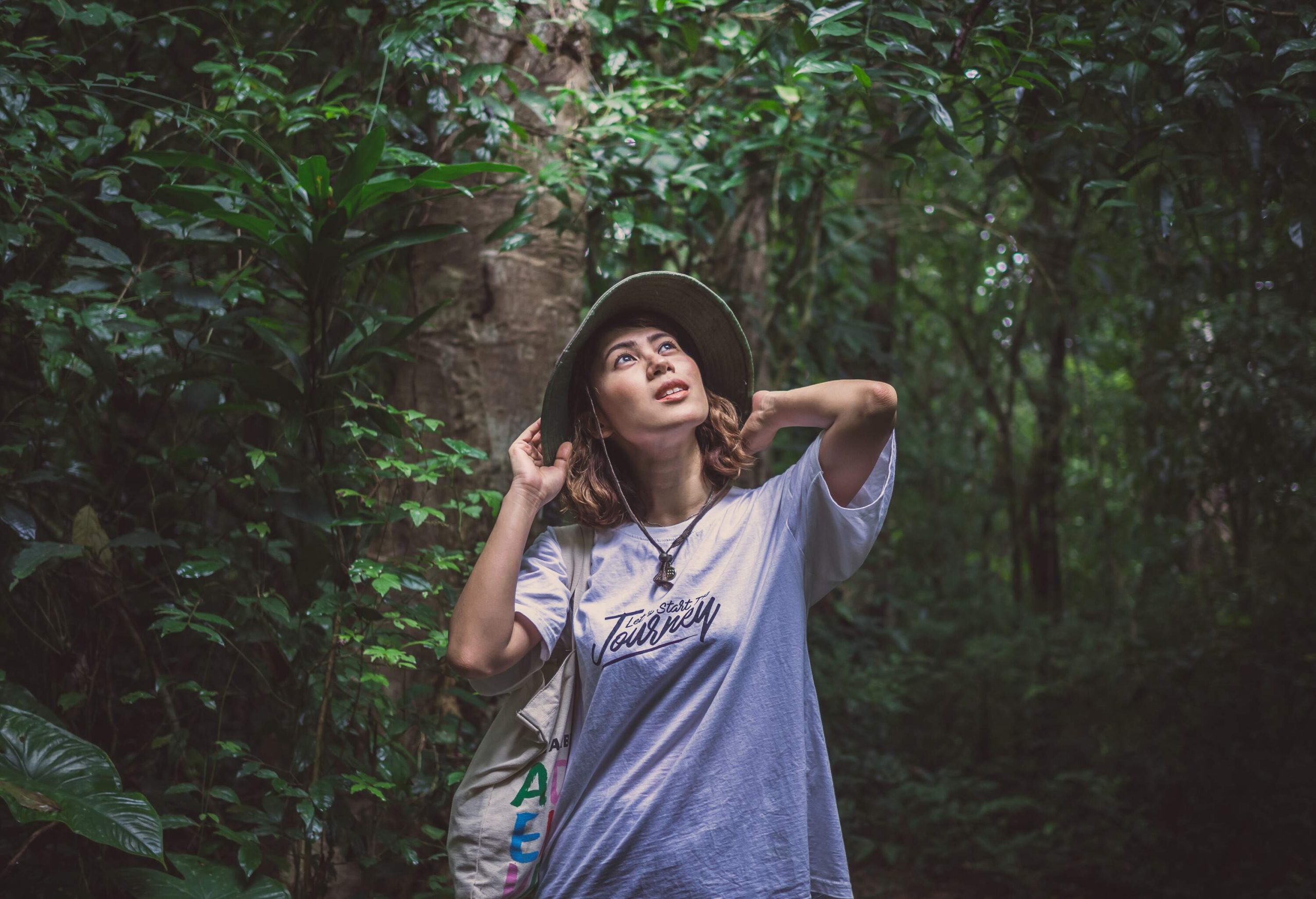 A woman wearing a white shirt, a green bucket hat, and a canvas tote bag looking at a green forest.