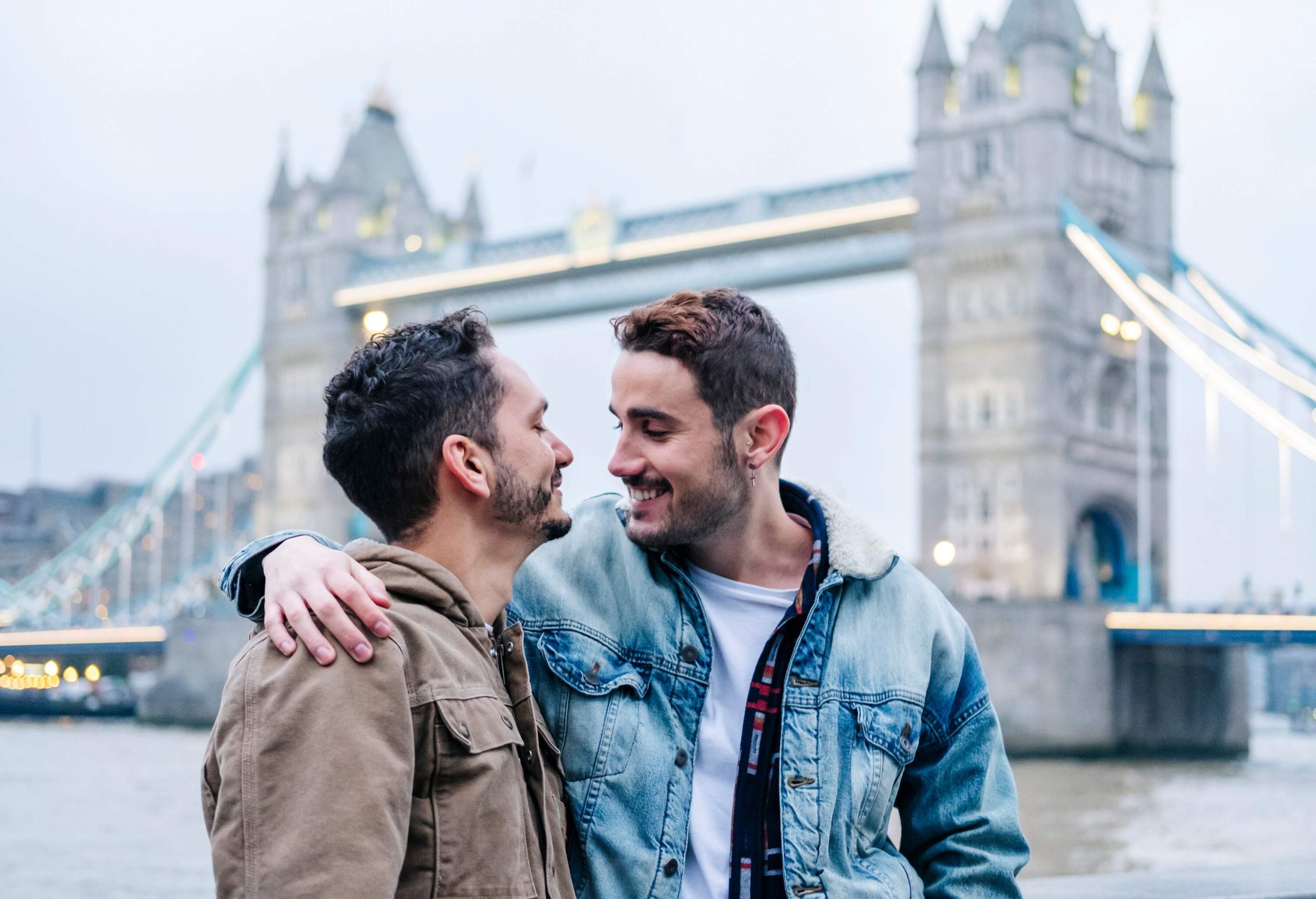Two male friends side-hugging against the London Tower bridge in the background.