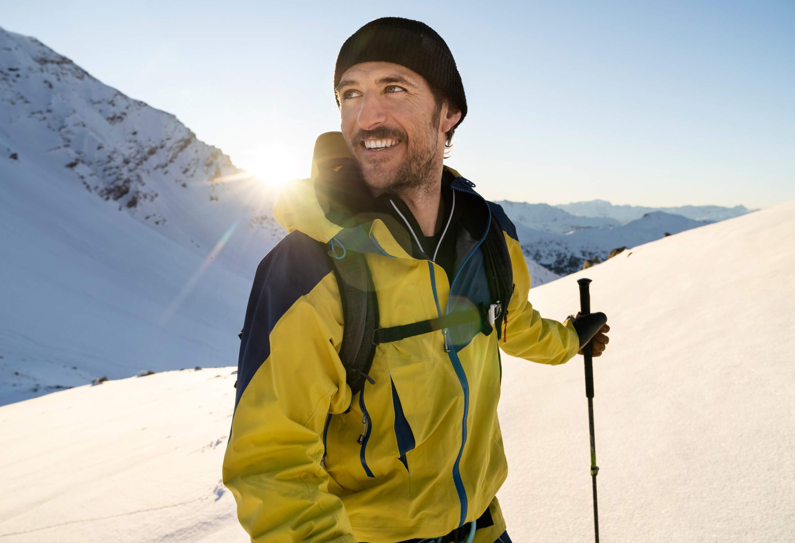 The sun shines behind a smiling man in a yellow jacket holding a ski pole.