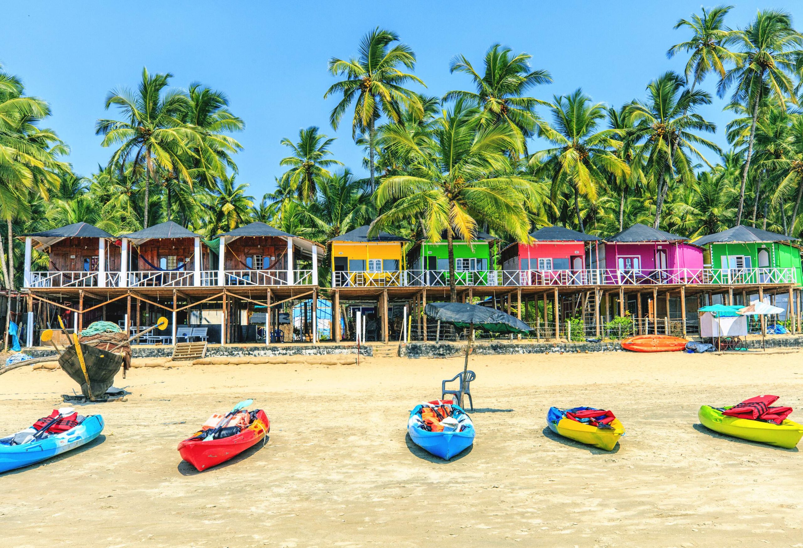 A row of multi-coloured elevated bungalows surrounded by lush palm trees facing the beach with tiny boats.
