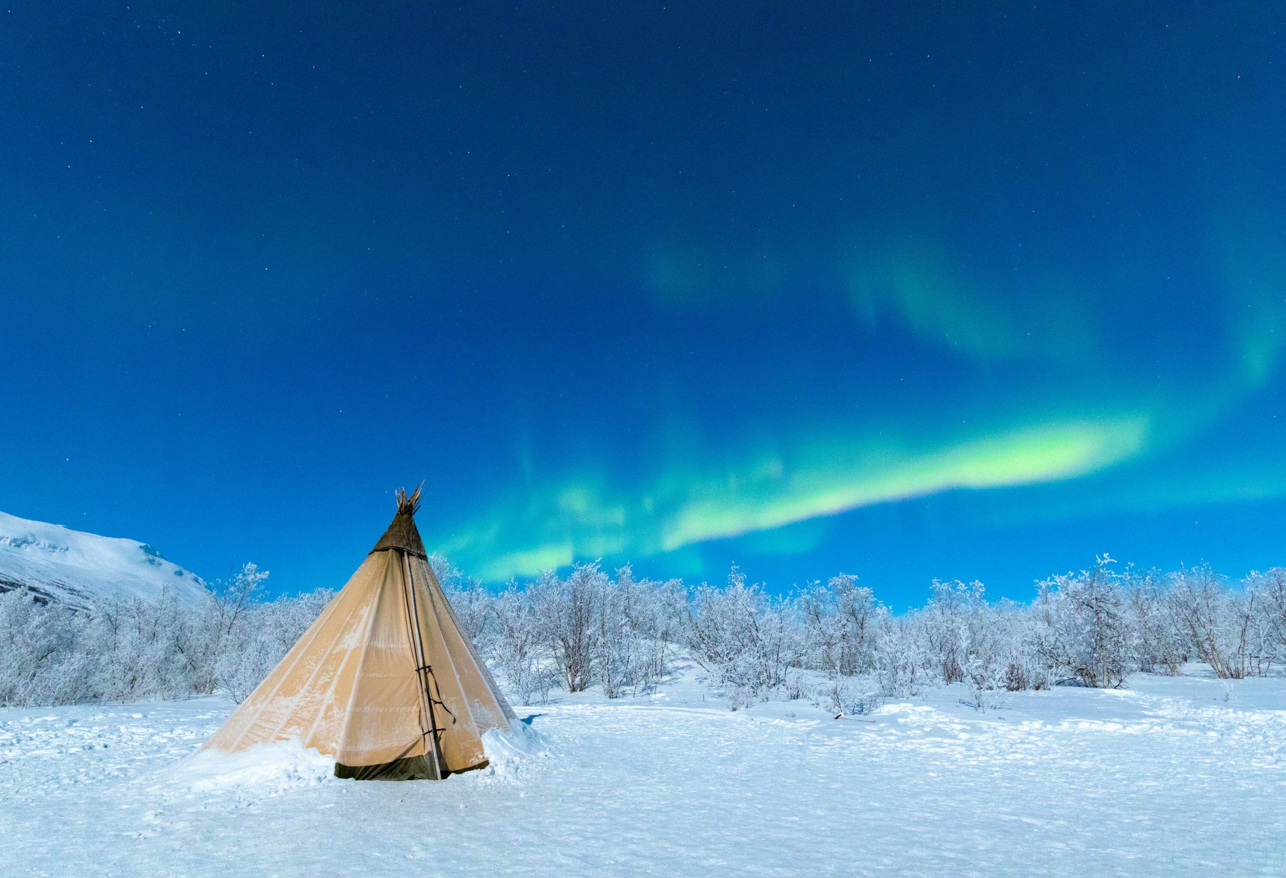 A Lavvu tent pitched on a snowfield with the northern lights swirling across the blue sky.