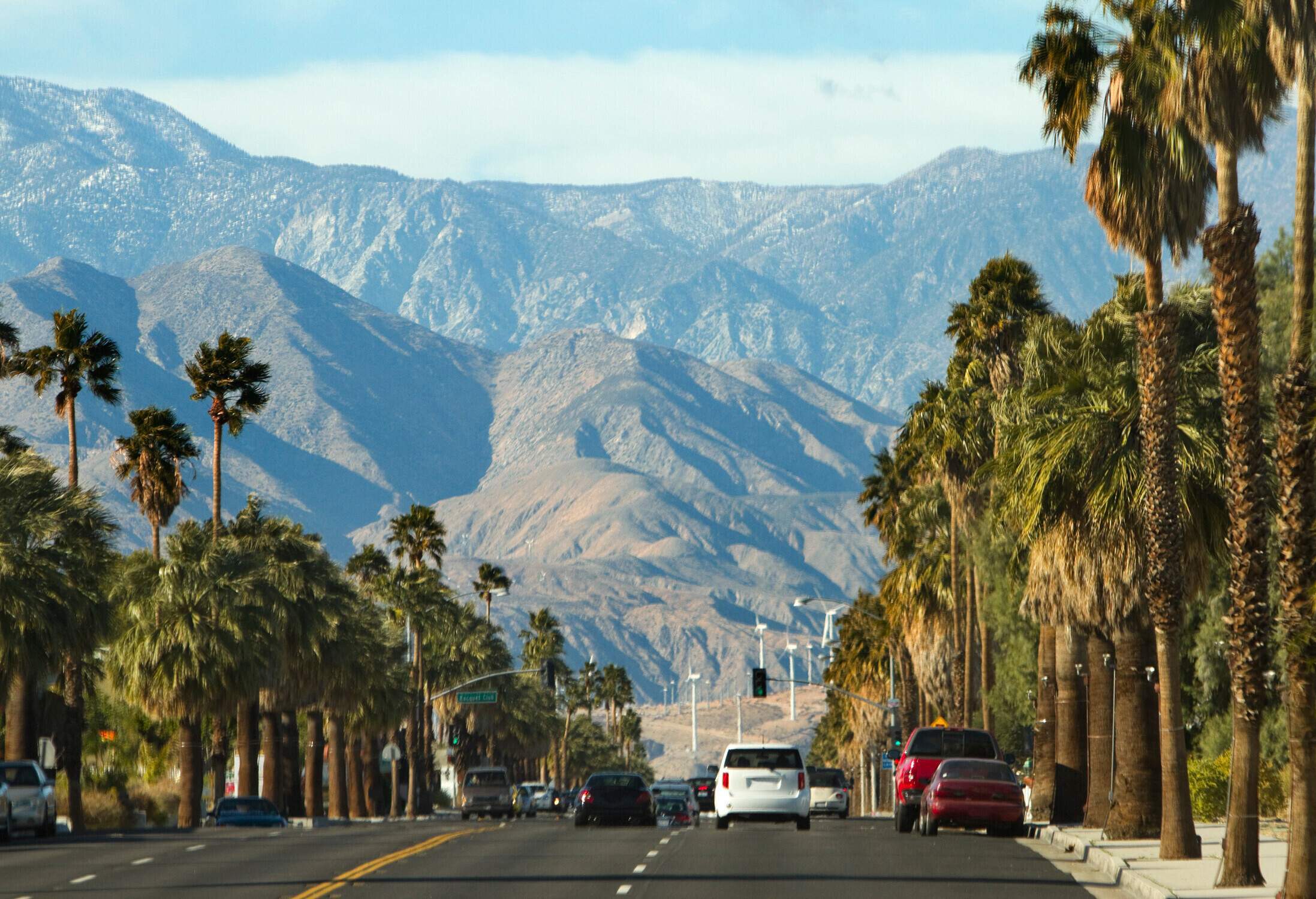 A scenic road, bustling with cars, is adorned with palm-lined sidewalks, while a majestic mountain range looms in the distance.