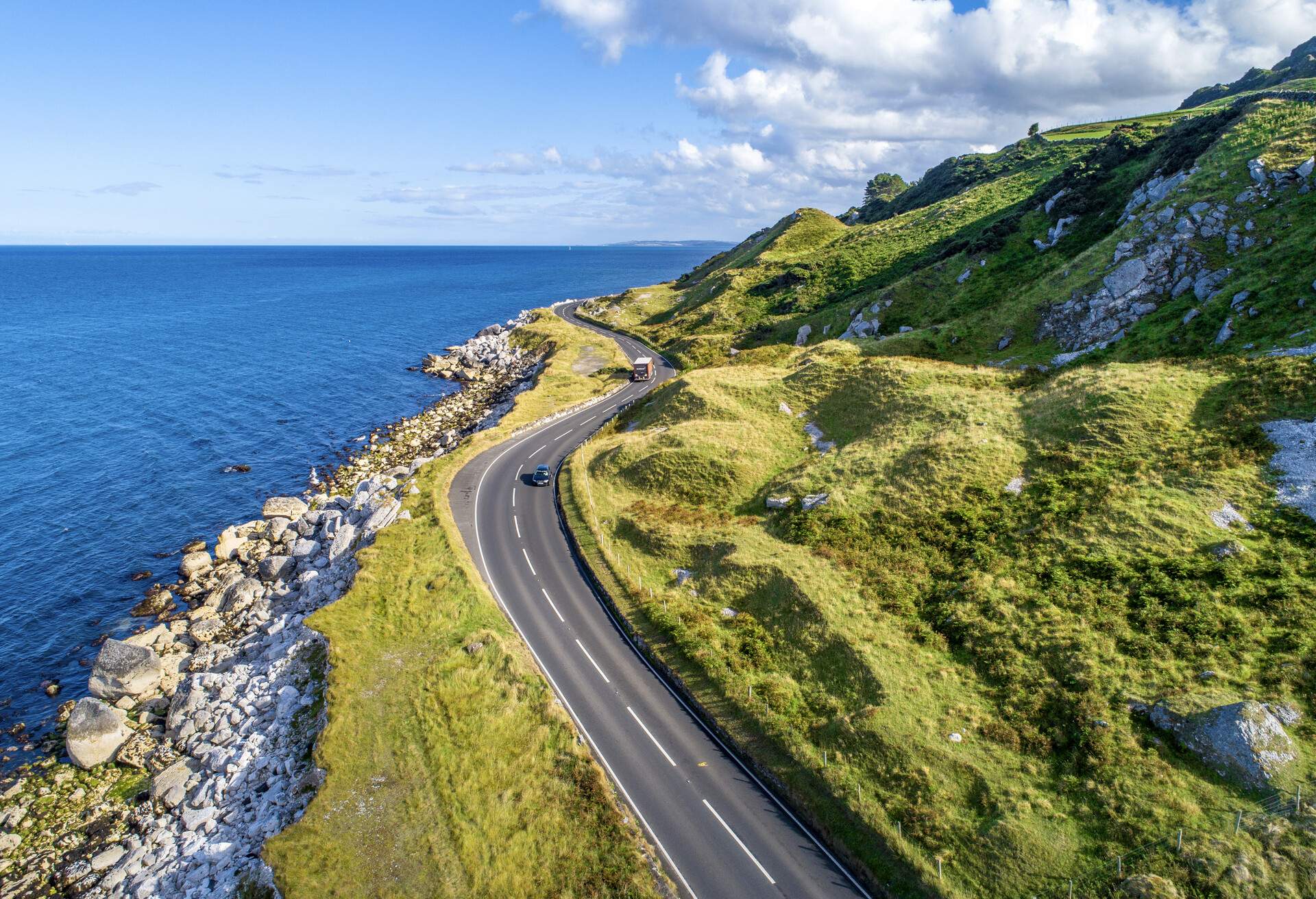 Cars passing by a coastal road beside the rocky hills covered in green foliage.