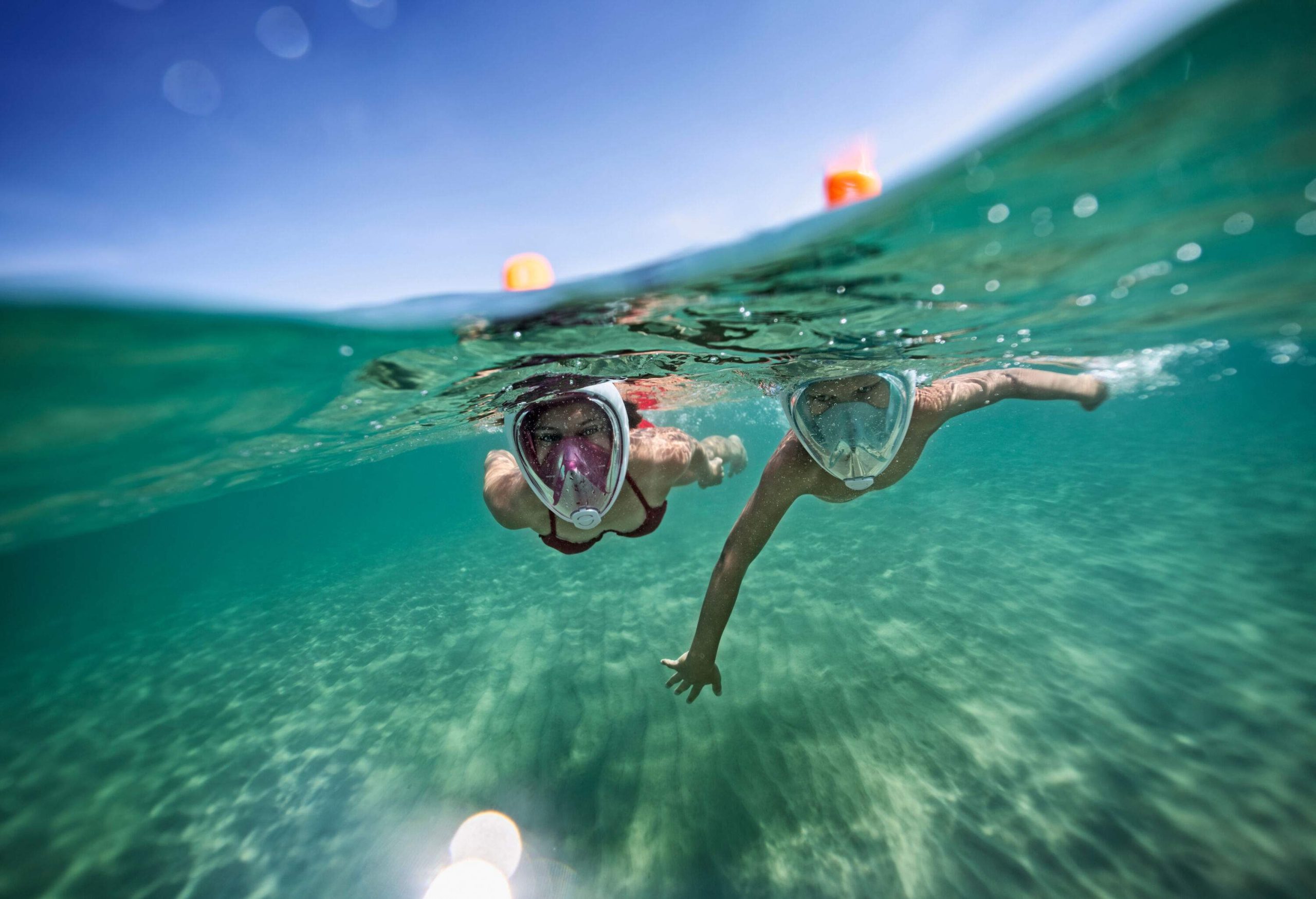 A boy and a girl in full-face snorkel masks swimming below the sea's surface under the cloudless sky.