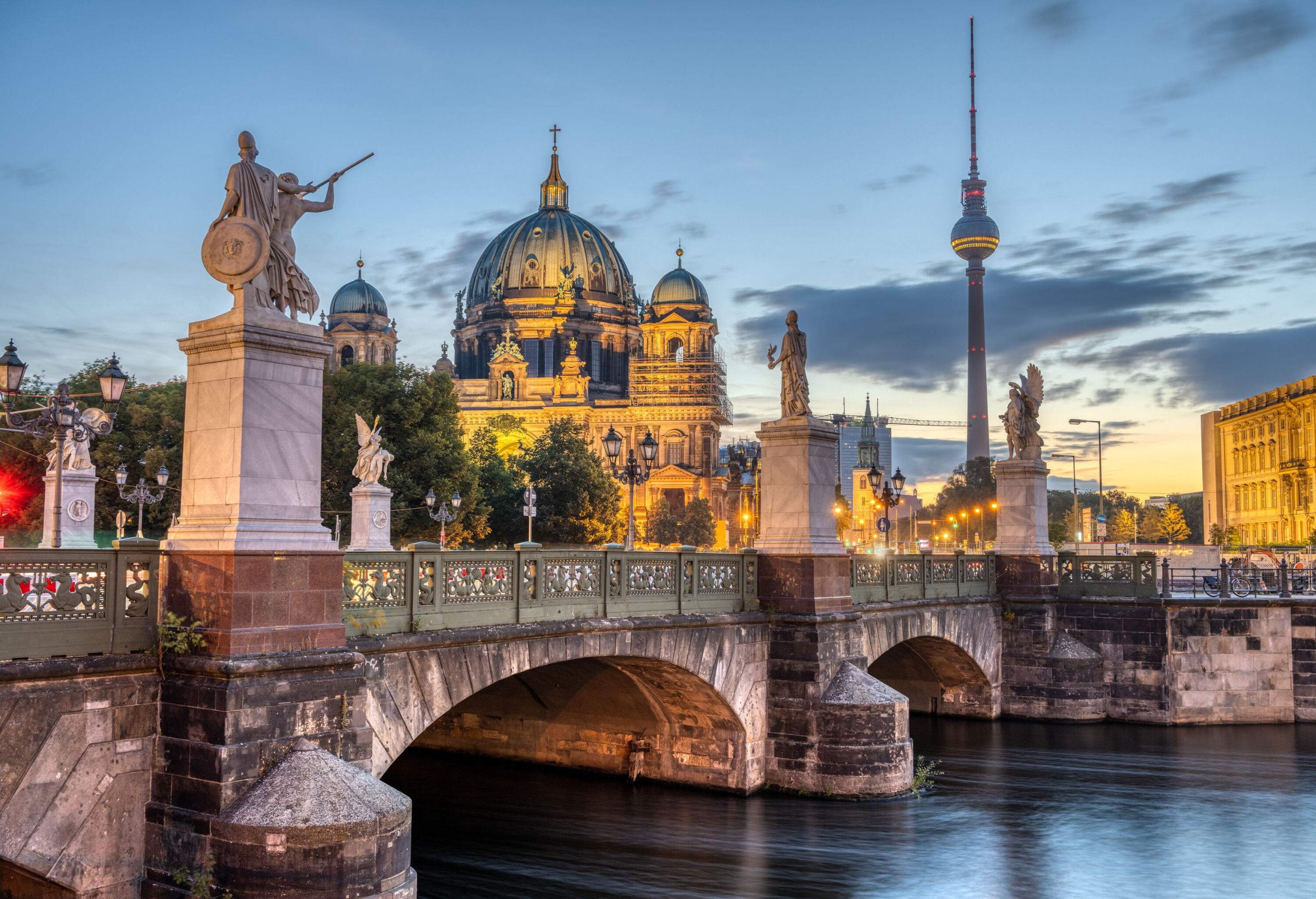 A sandstone bridge lined with sculptures across a river that offer views of a domed palace and a distant television tower.