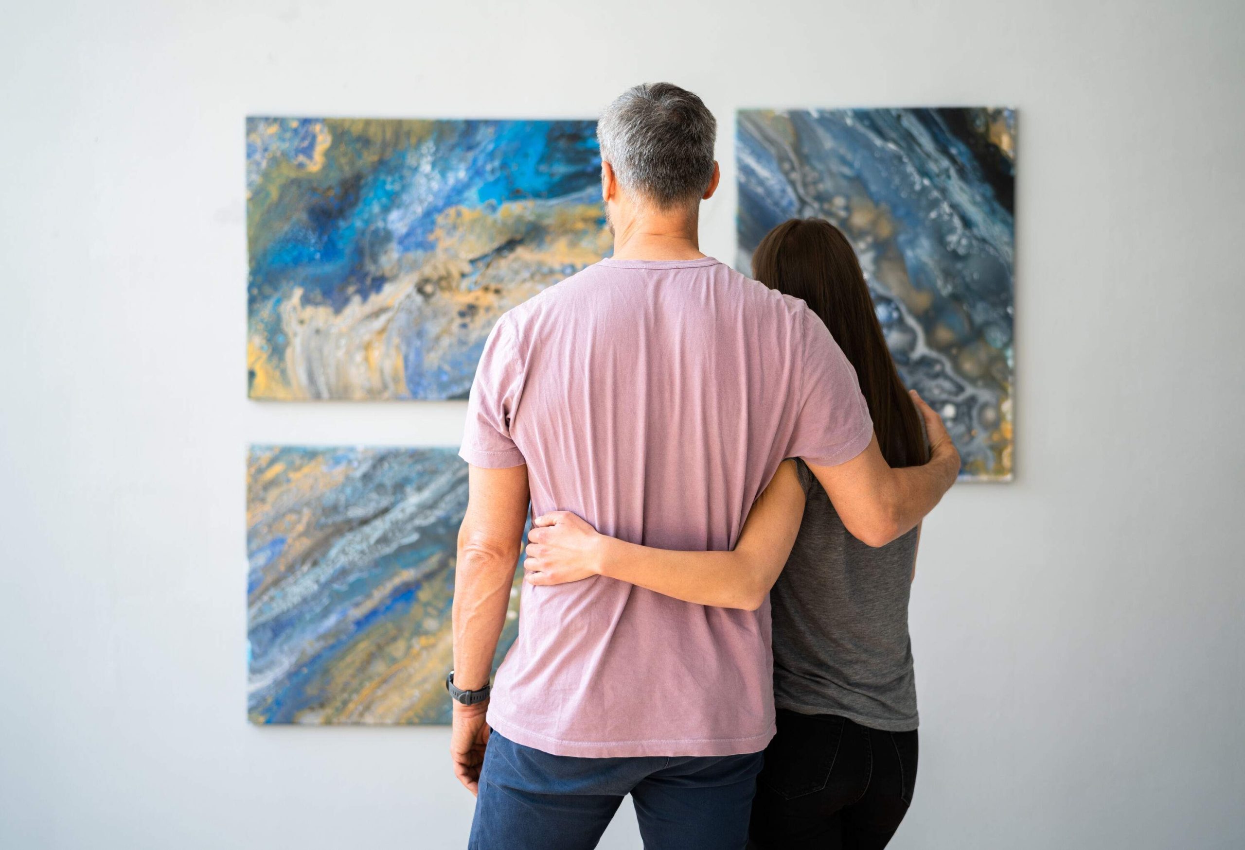 A man and a woman with arms around each other looking at paintings on the wall.