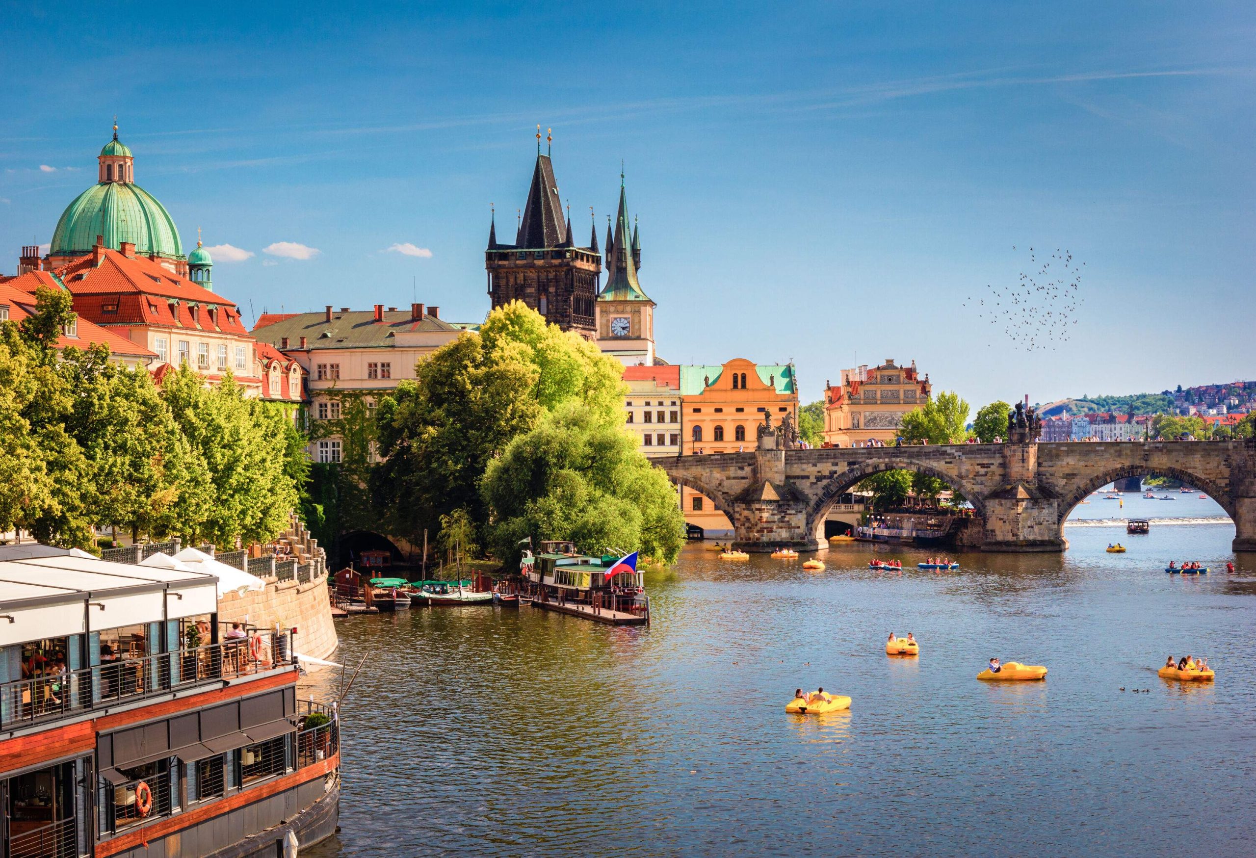 The iconic Charles Bridge extends over a river teeming with people enjoying a day out on yellow paddle boats, with passenger boats moored on the side, all set against the grandeur of Prague Castle looming in the background.