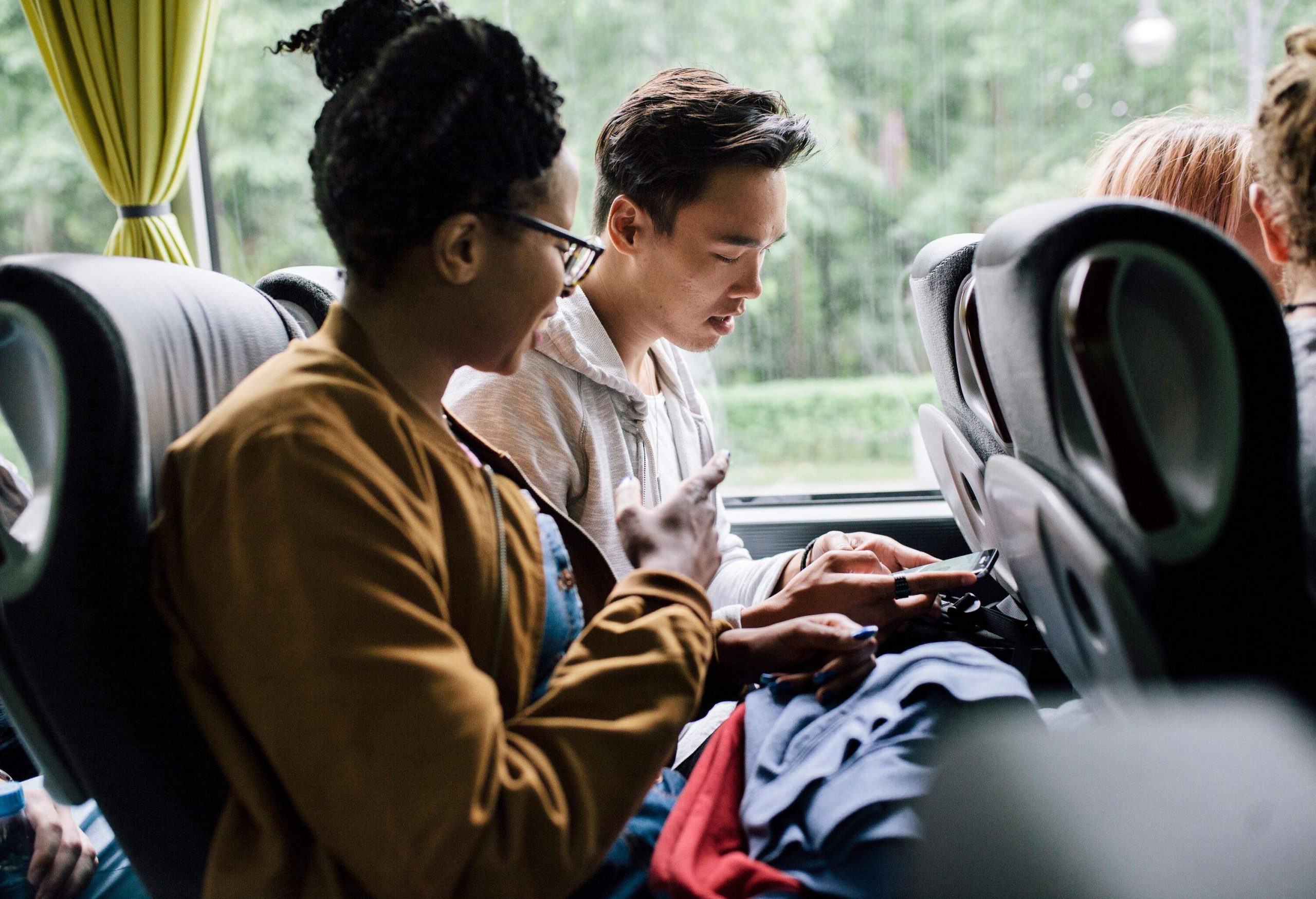 Two friends sit and have a conversation inside a bus, with one using his phone.