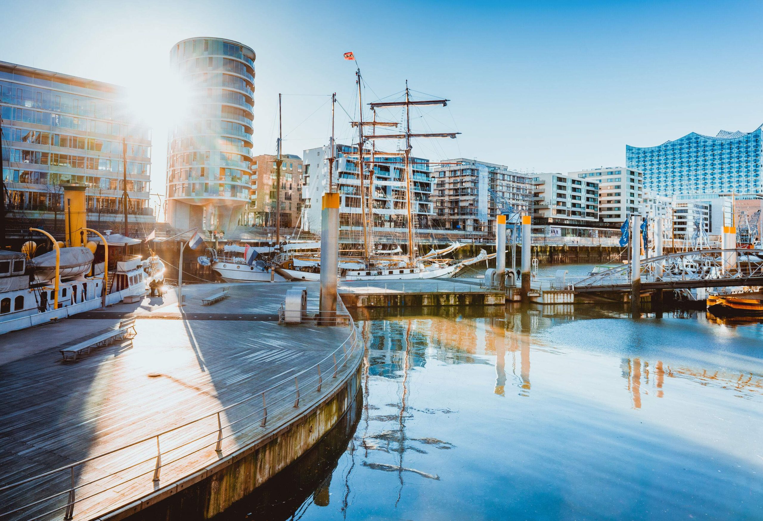 A promenade on a port with anchored boats and views of the city skyline.