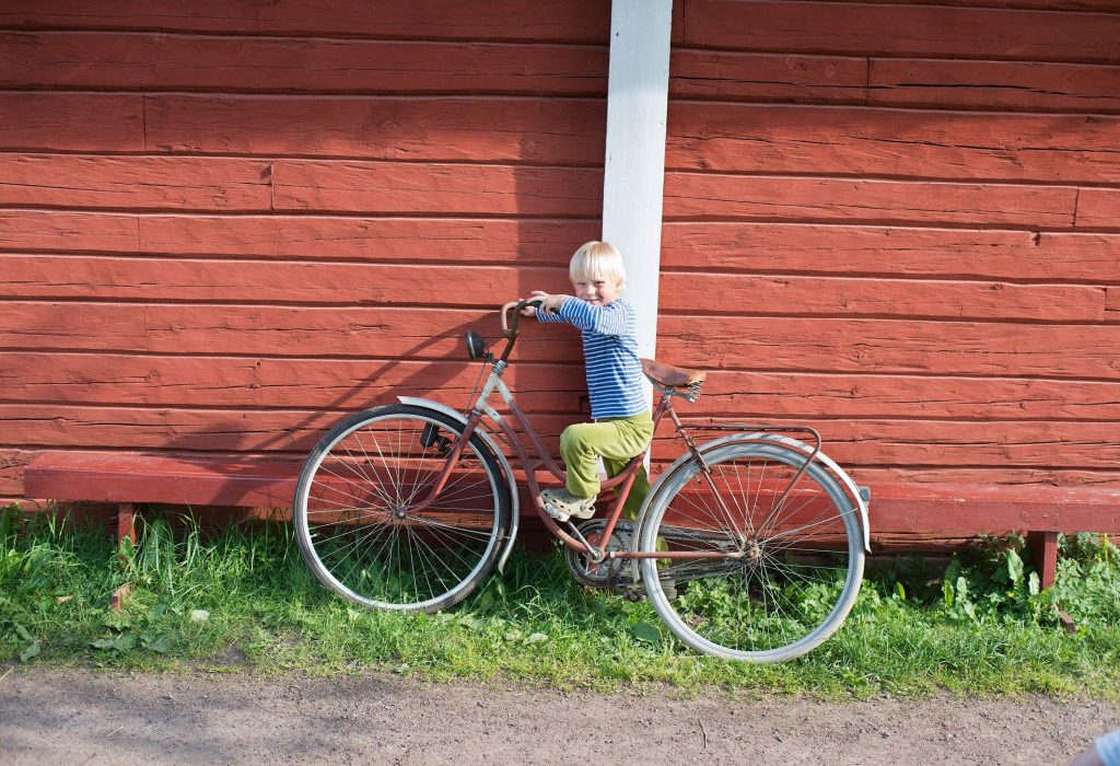 A youngster wearing a striped shirt rides a bicycle that is leaning against a wooden wall.