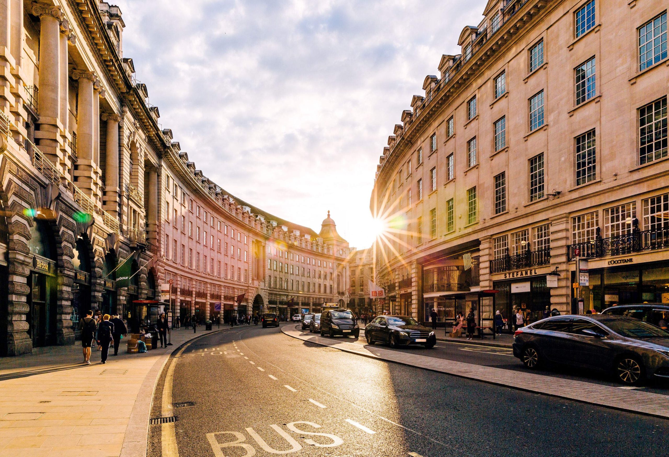 The sun's rays shine through a curved bustling street between the rows of buildings. 