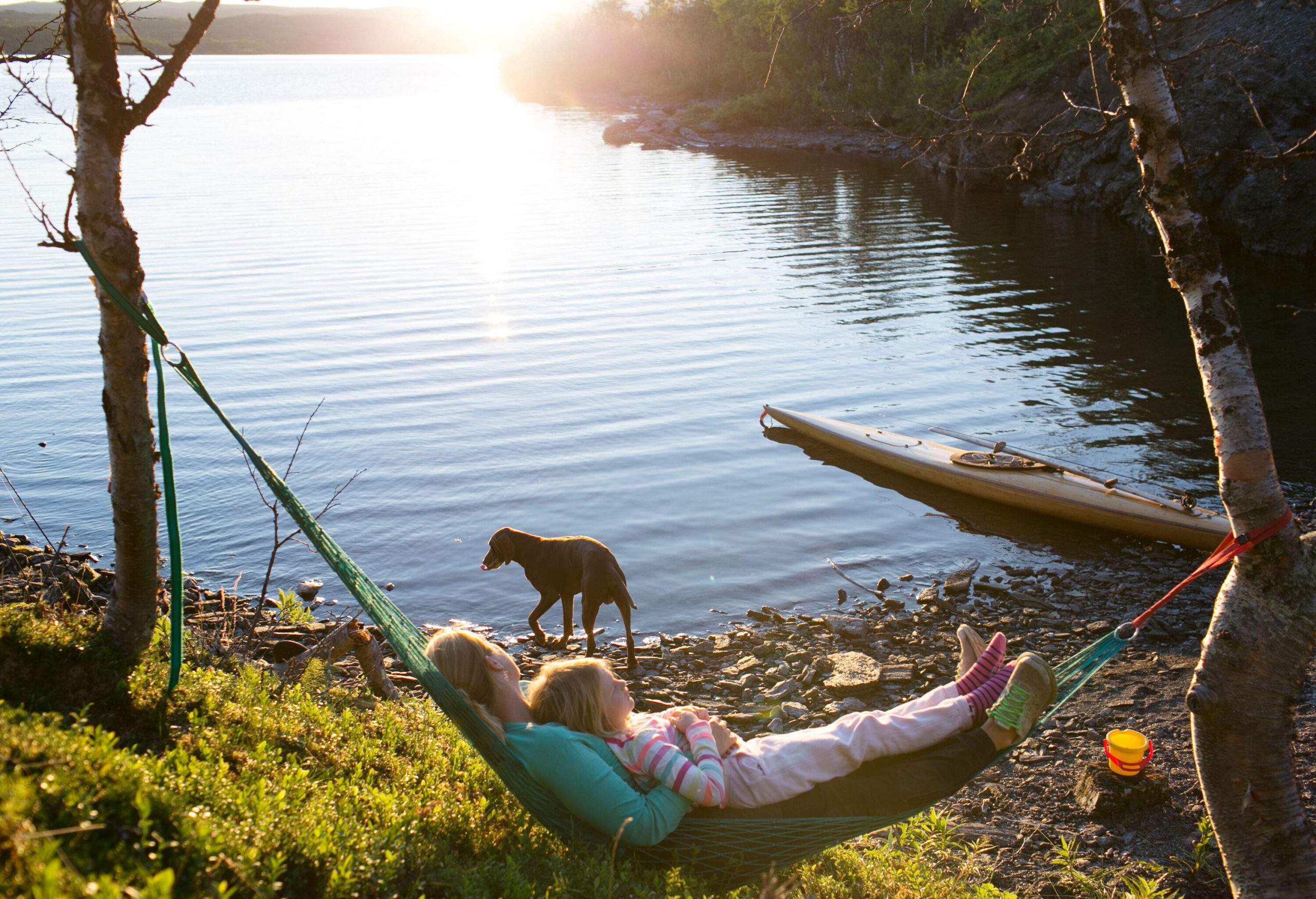 A mother and daughter are on a hammock with their dog by the lake as they enjoy the sunset.