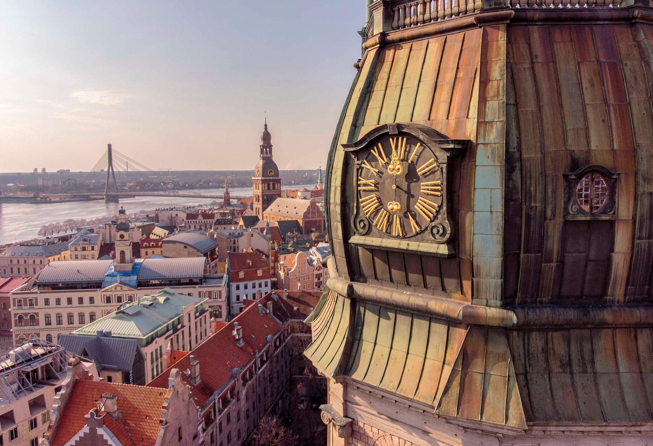 A close-up view of a clock on a church tower overlooking the river along the compact buildings and spanned by a cabled stayed bridge.