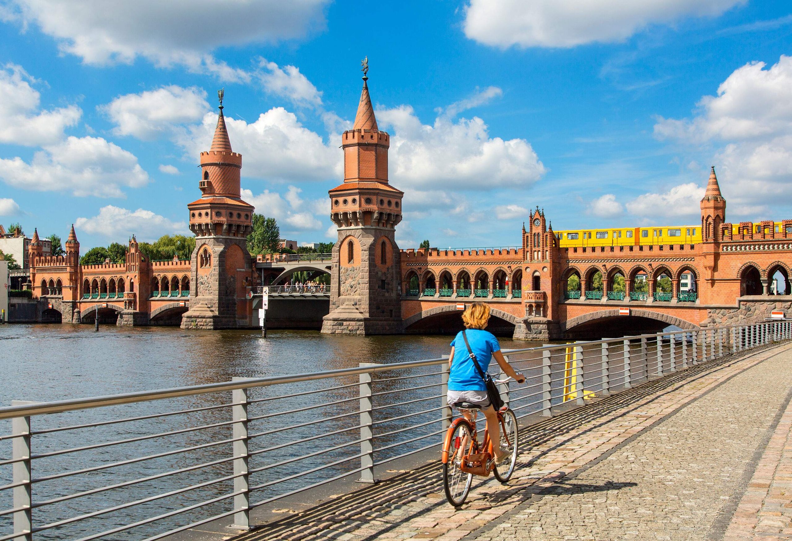 A person riding a bike across the promenade by the river with a double-deck bridge.