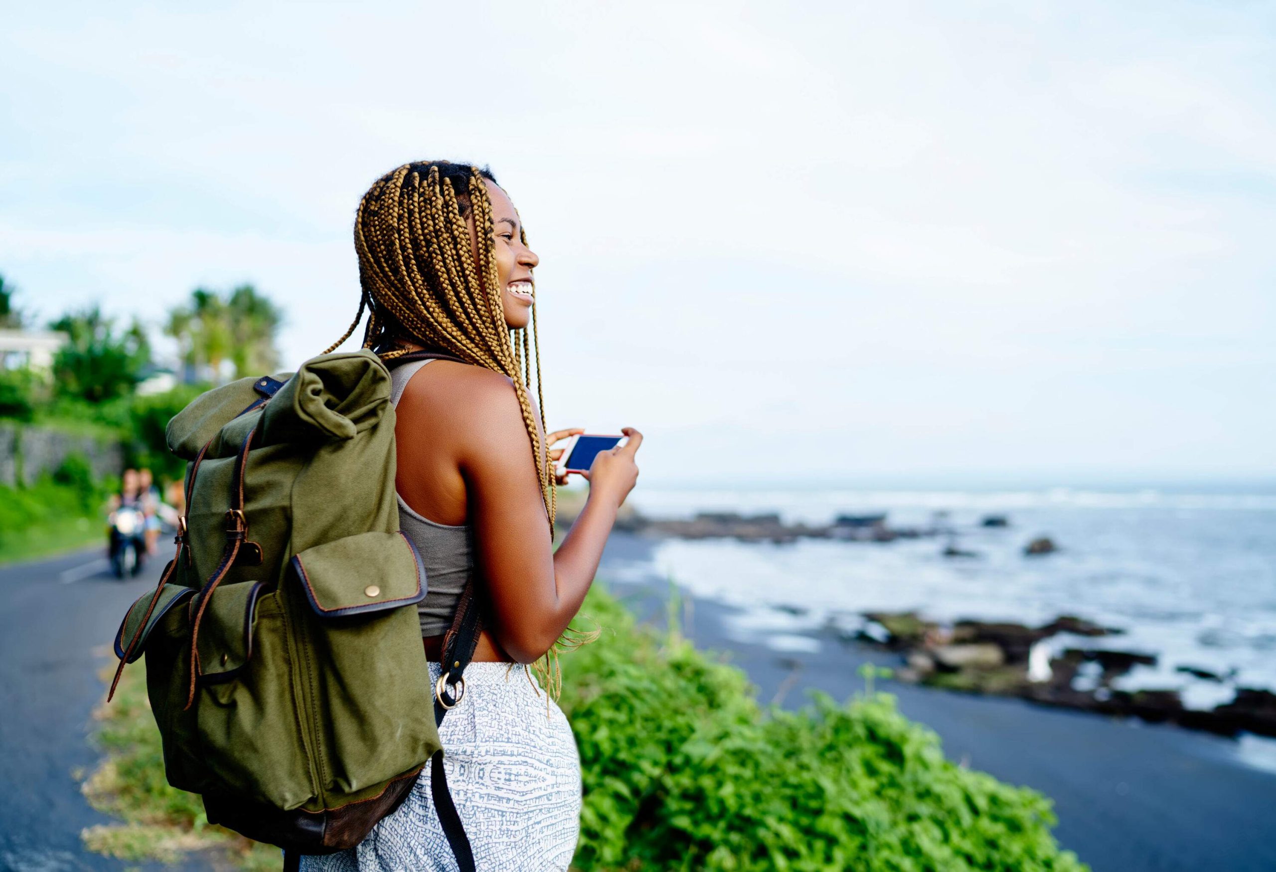 A woman with box braids and a canvas backpack standing on the shore with a smartphone.