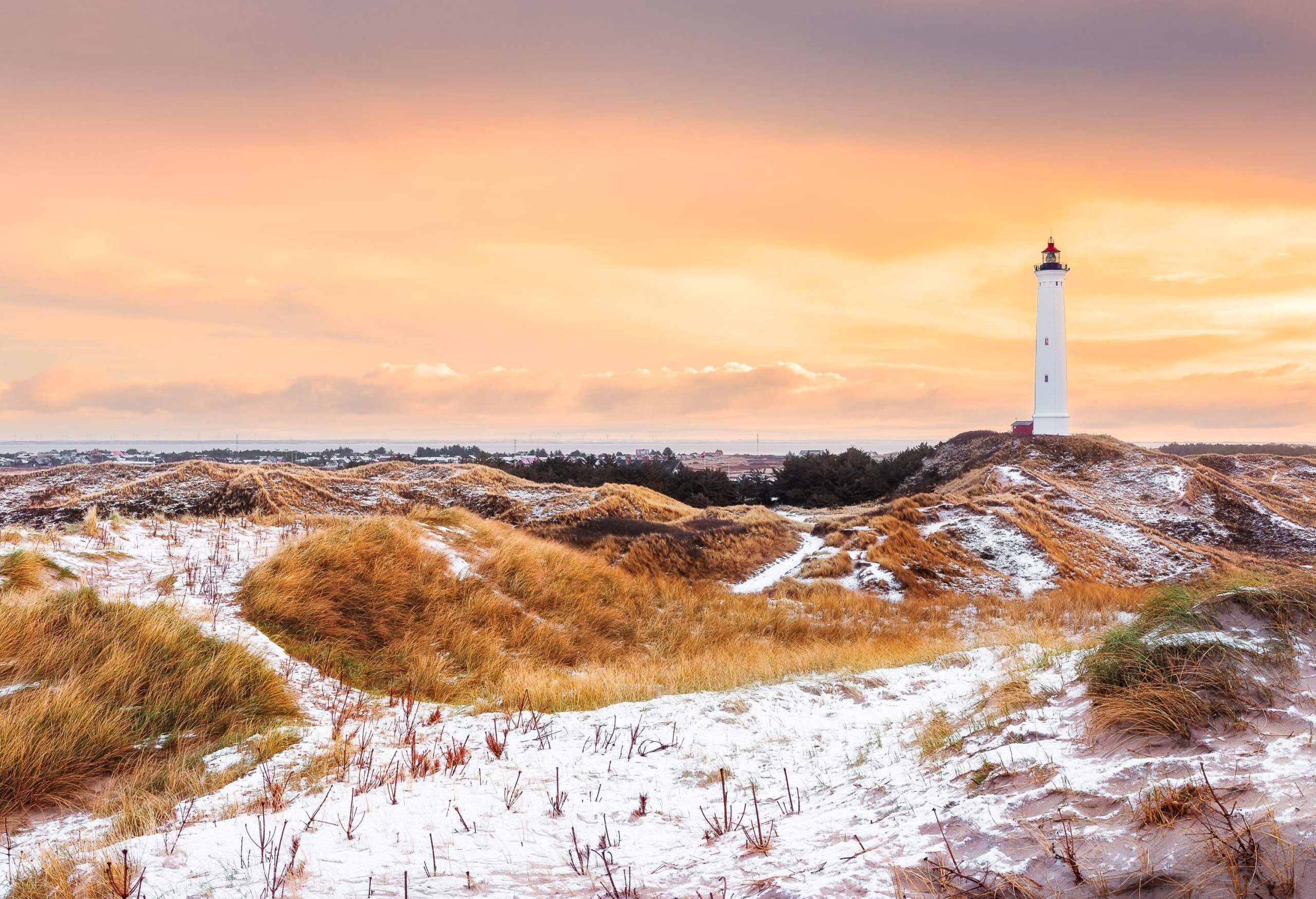 A white lighthouse stands on a snow-covered meadow under an orange sky.