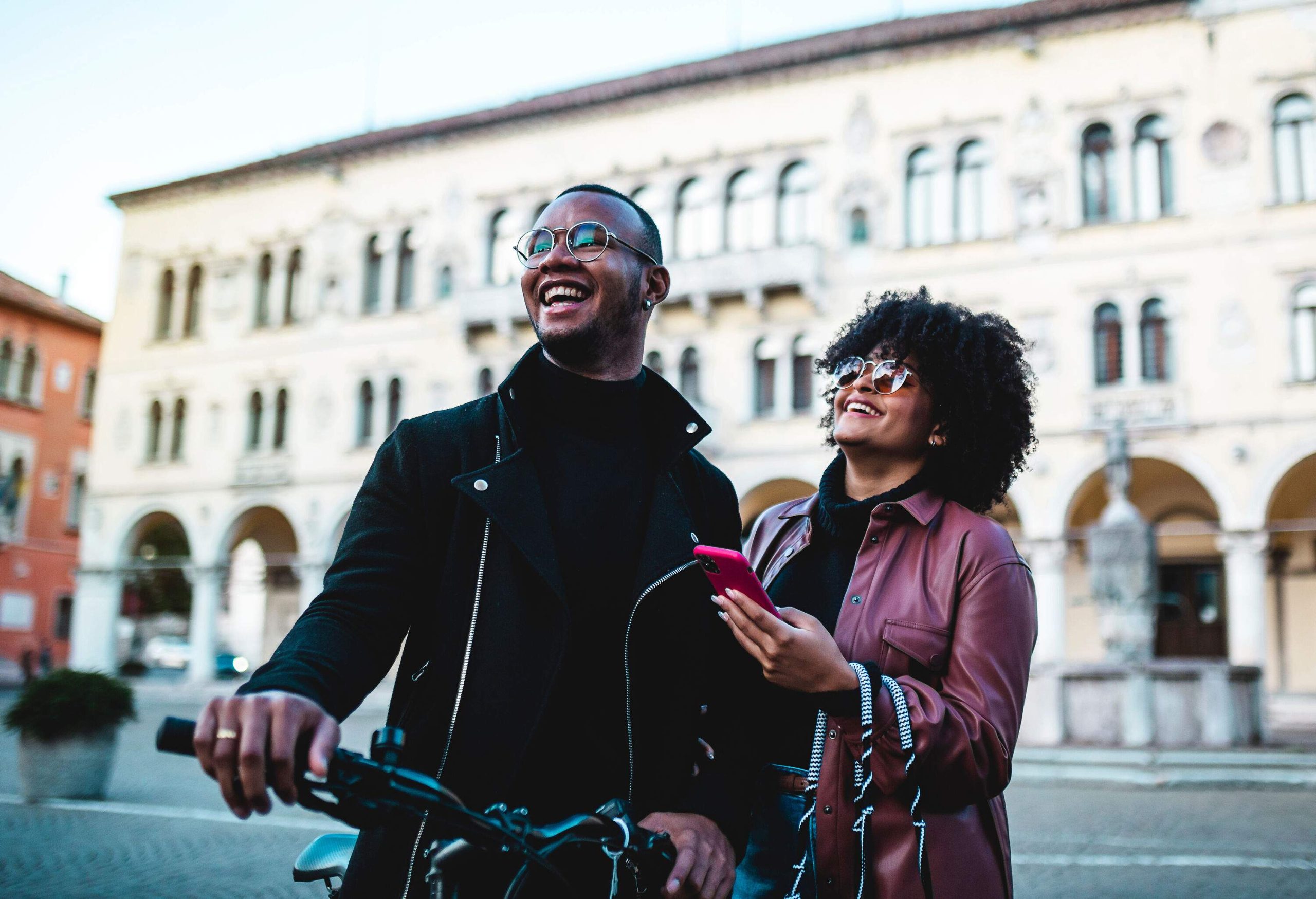 A man with his bike laughs with his girlfriend on the street next to a building.