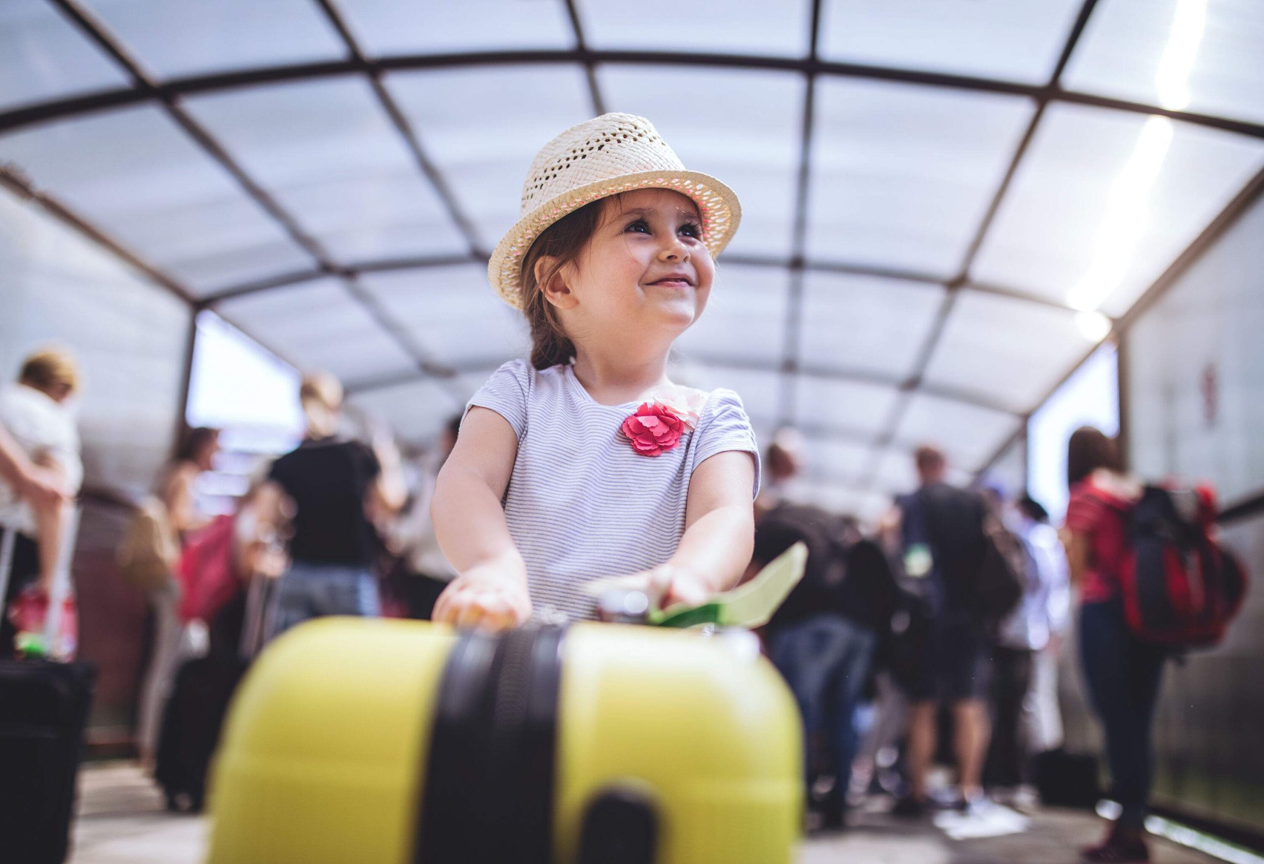 A cute little girl holds onto a yellow suitcase against the busy crowd in the background.