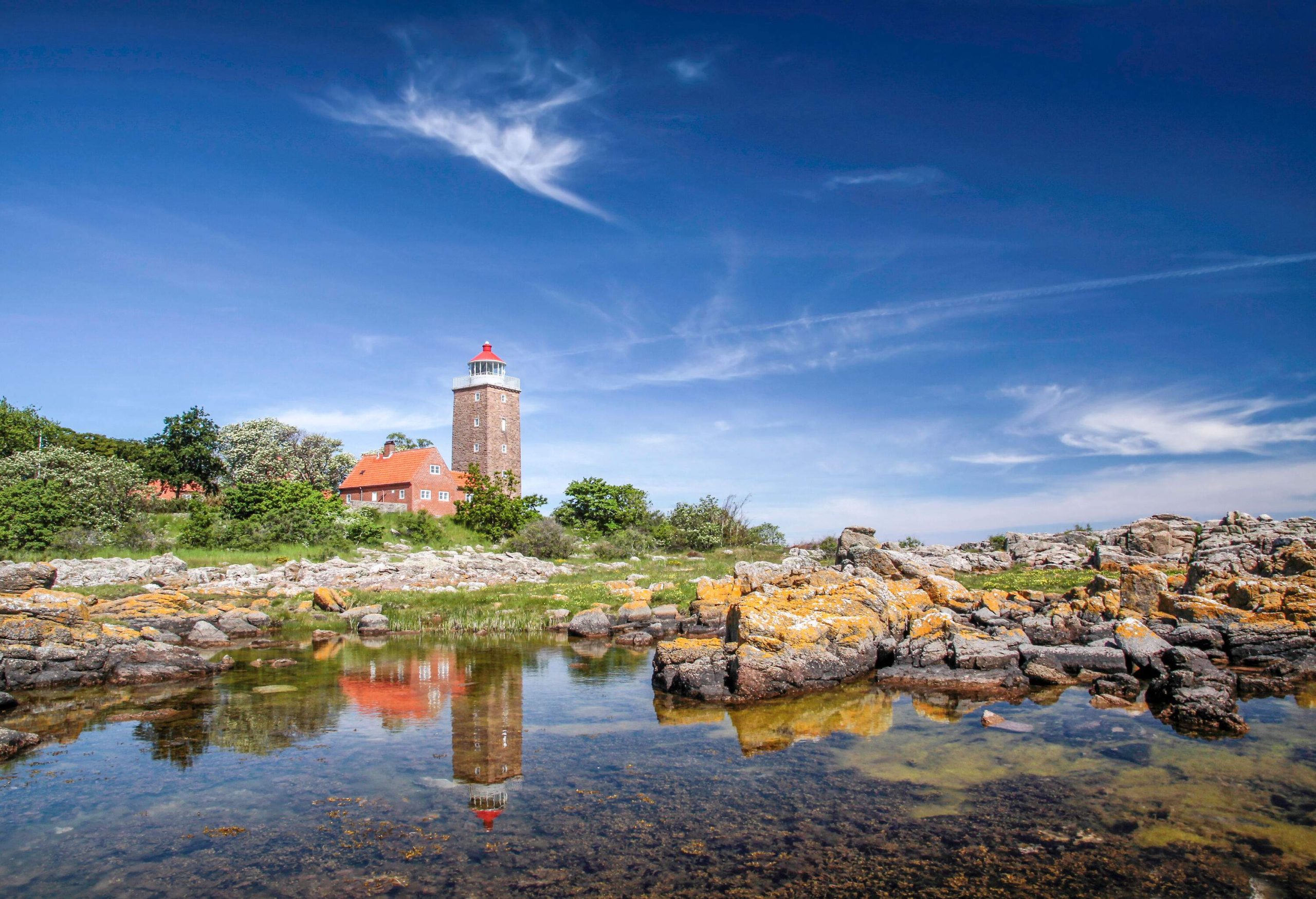 A square-shaped lighthouse and keeper's house reflecting on shallow waters with a rocky shore.