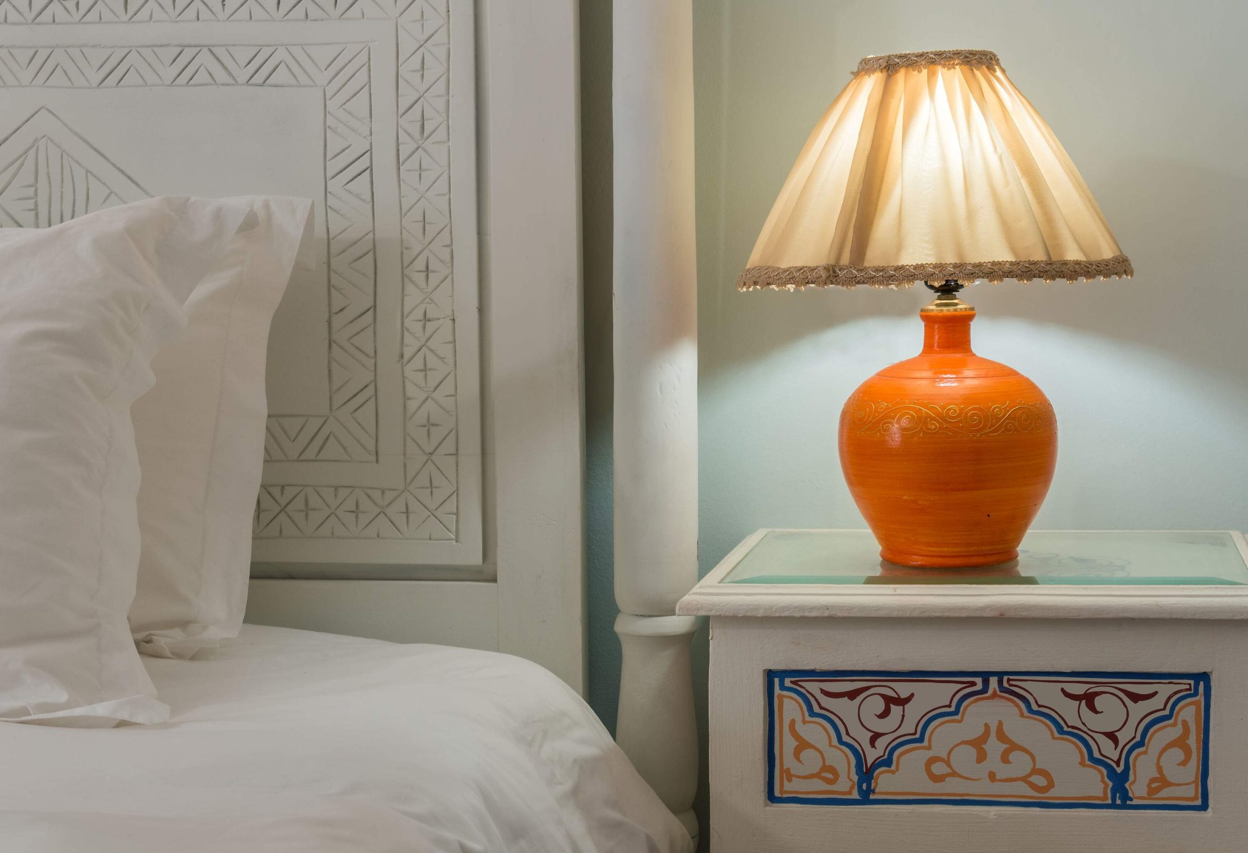 A lit lampshade on top of a small table beside a white bed.