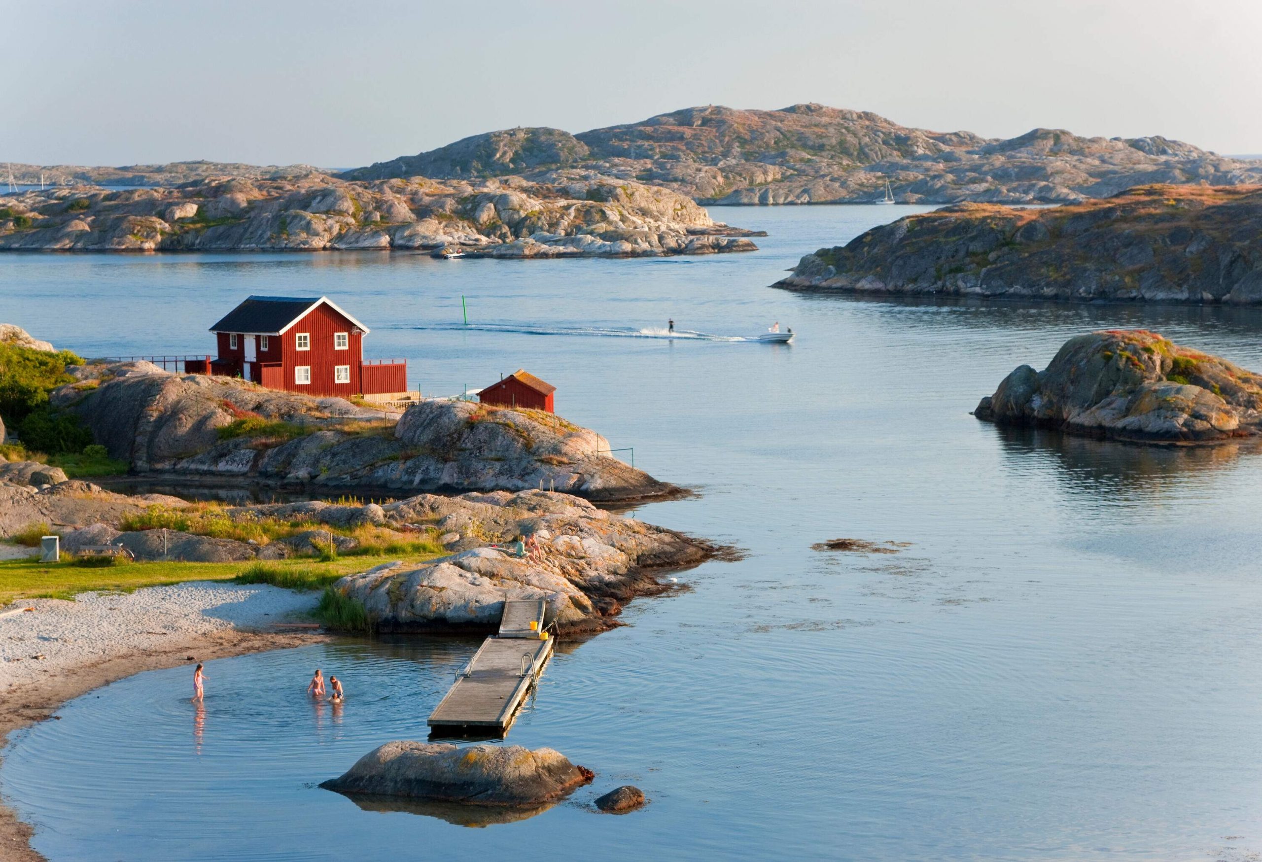 A family of four swims alongside a boardwalk while another group skurfing in the middle of rock islands with a red Swedish house.