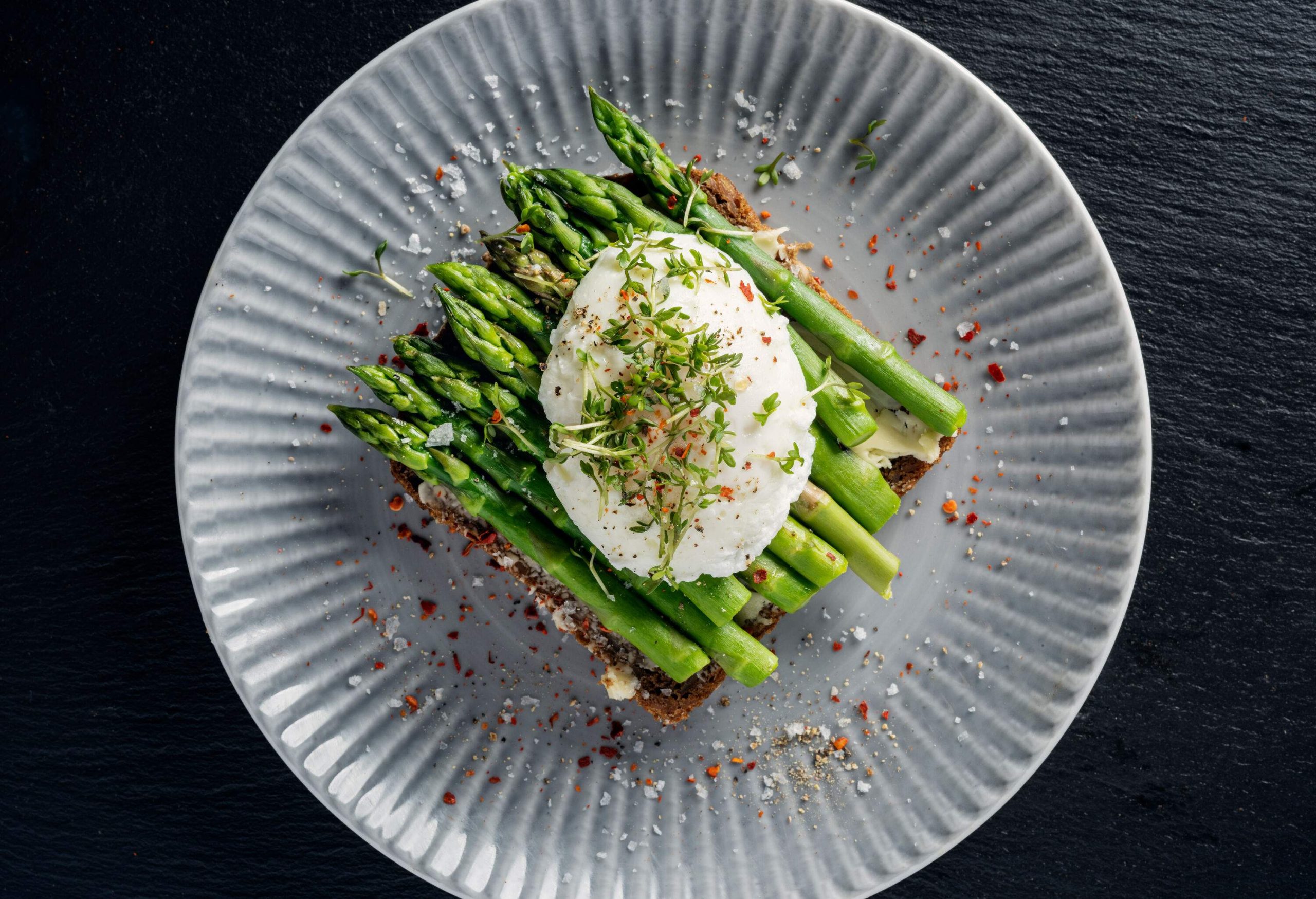 An open-faced sandwich topped with green asparagus and a poached egg garnished on a plate.