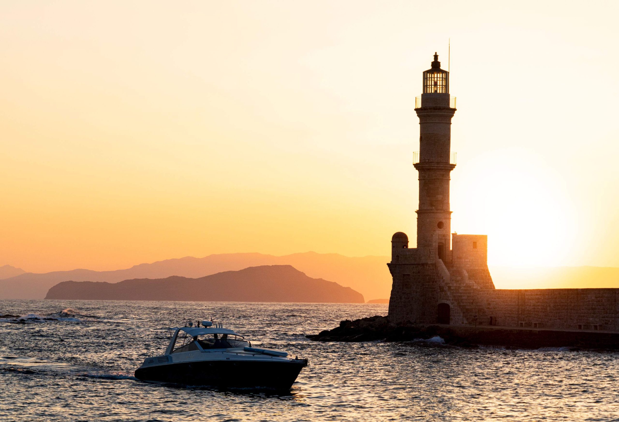 Silhouette of a lighthouse near a cruising yacht against the backdrop of the glaring sun.