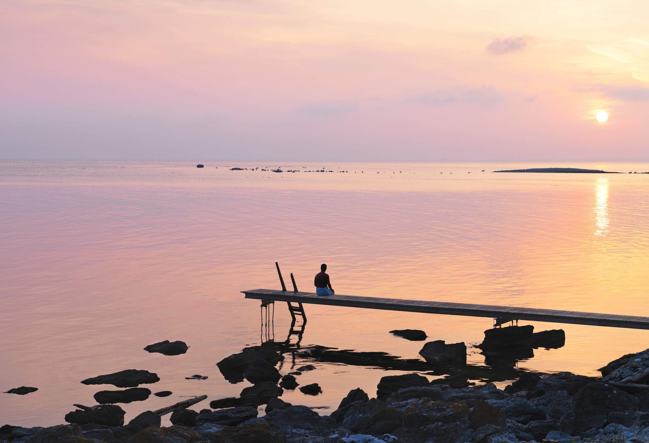 Silhouette of a person sitting on a jetty of a rocky beach while gazing at the sunset.