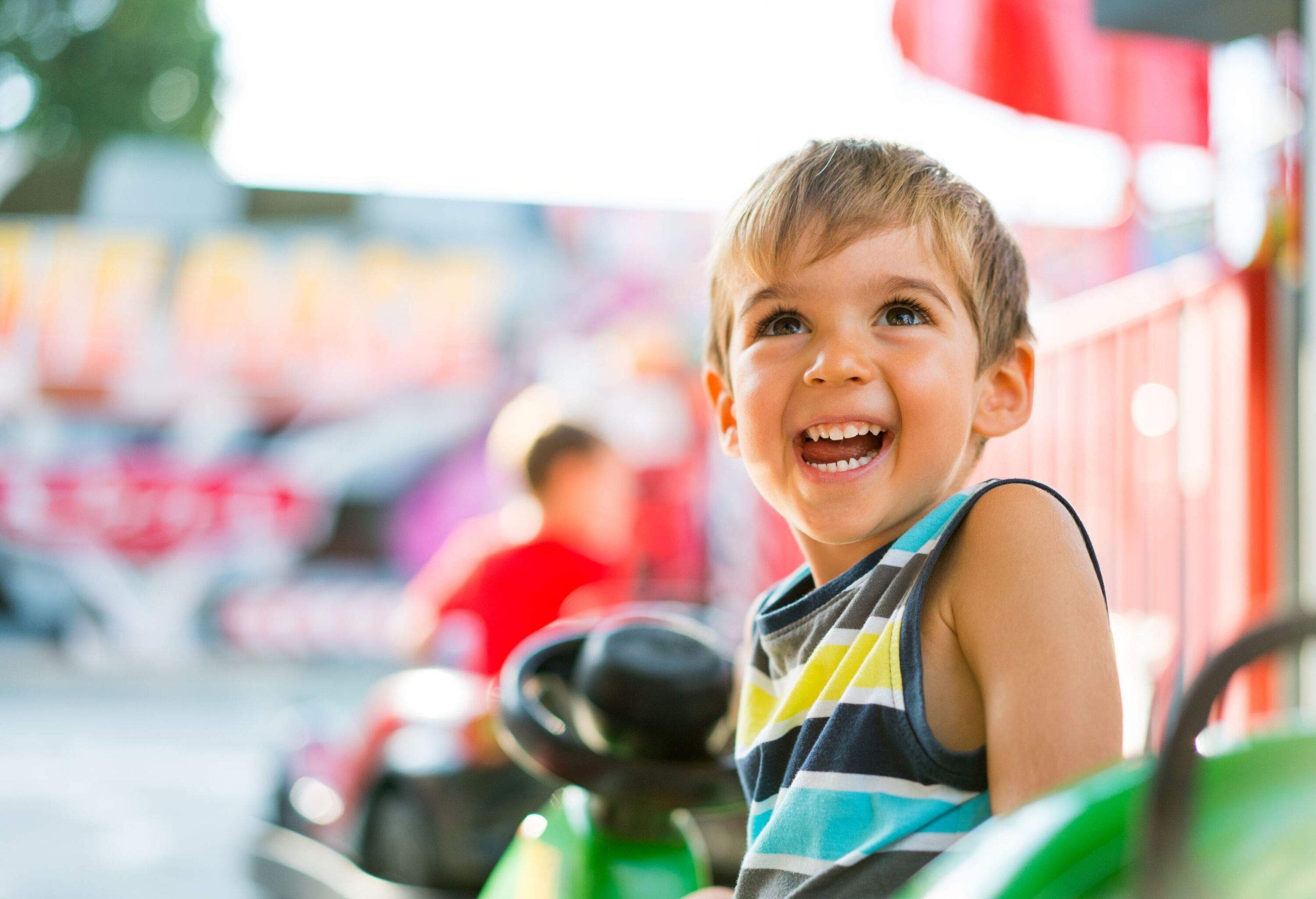 A young boy in colourful tank top smiles as he sits on a theme park ride.