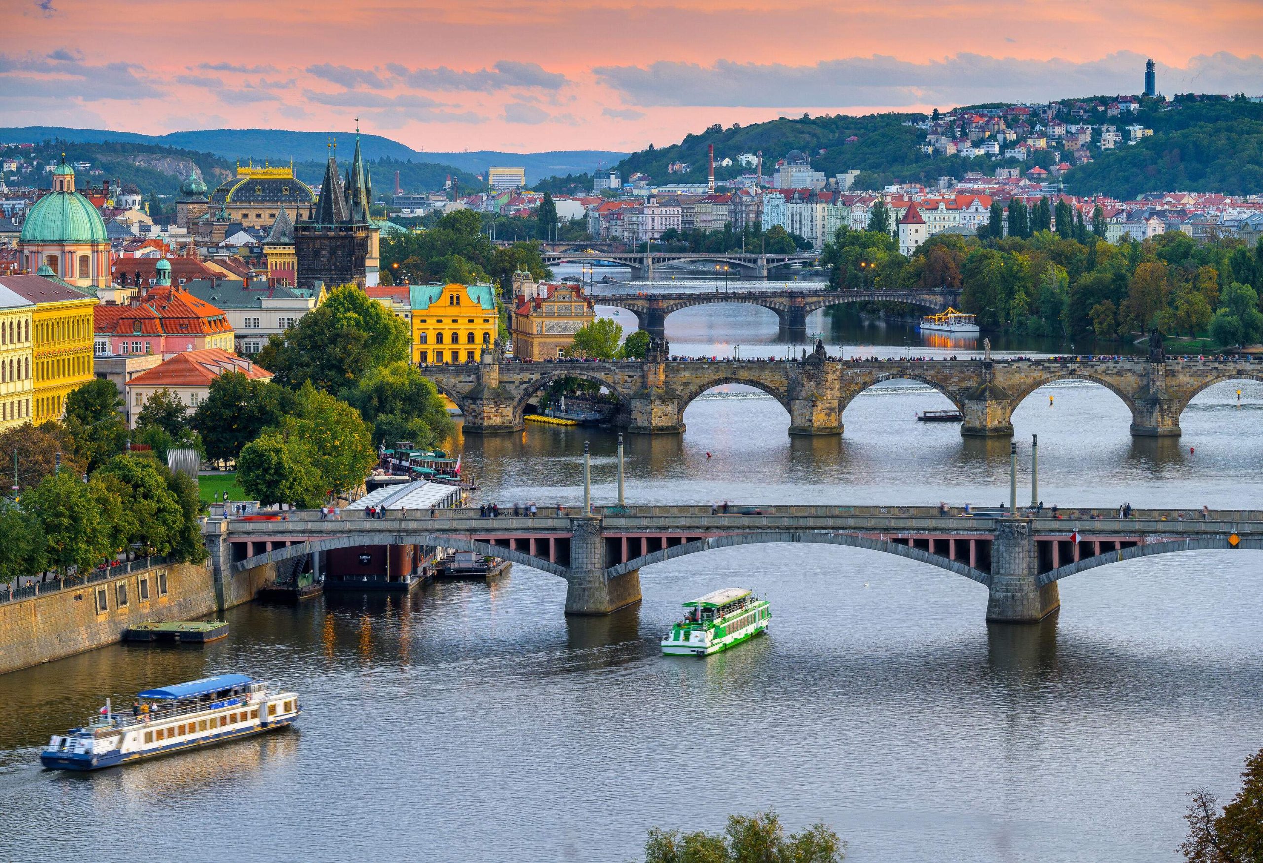 Multiple stone bridges gracefully span a river, framed by a charming town with vibrant, colourful buildings and lush trees, while buildings nestled on tree-clad slopes paint a serene picture in the distance.