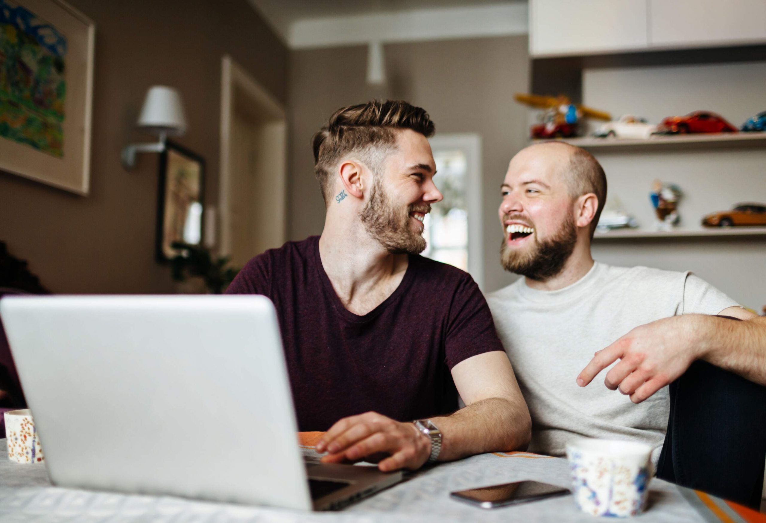 A happy gay couple is sitting in the dining area and holding a laptop in front of them.