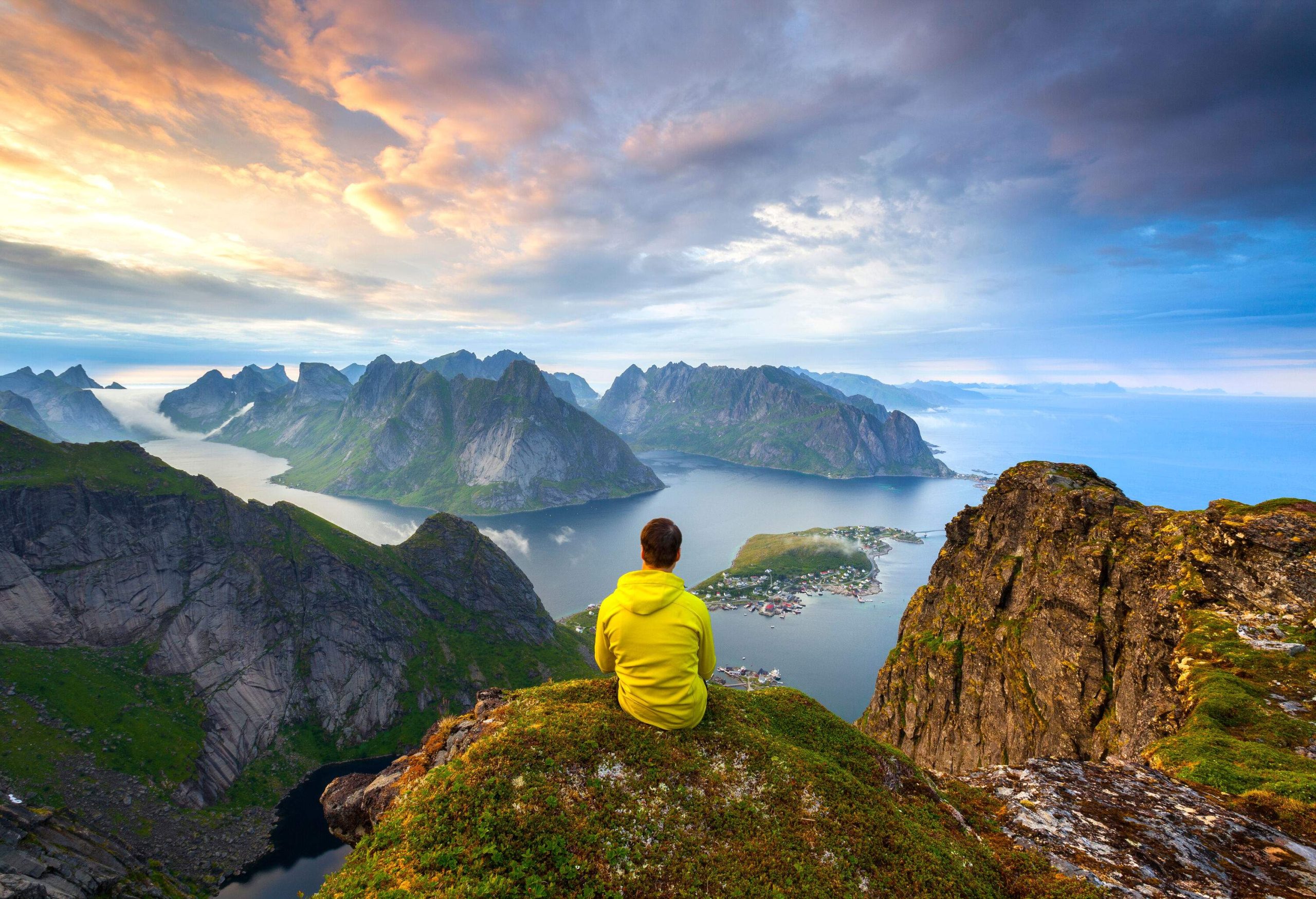 A man sits atop a rocky mountain with a spectacular view of steep rock islands emerging on the sea against the scenic cloudy sky.