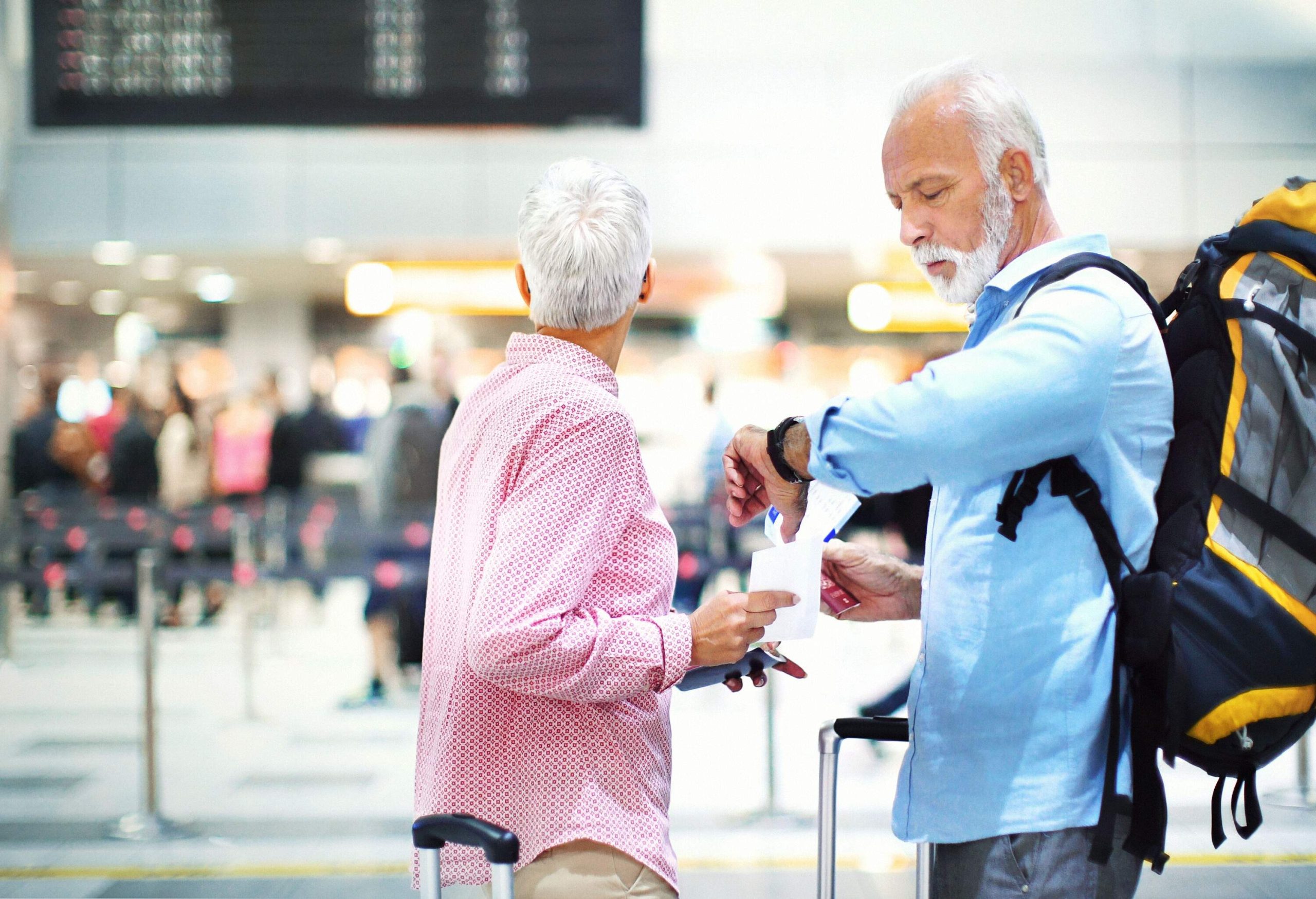 Closeup side view of senior couple at an airport waiting for their flight which has been delayed. They are both holding their passports and tickets. The lady is looking at the arrival departure board...