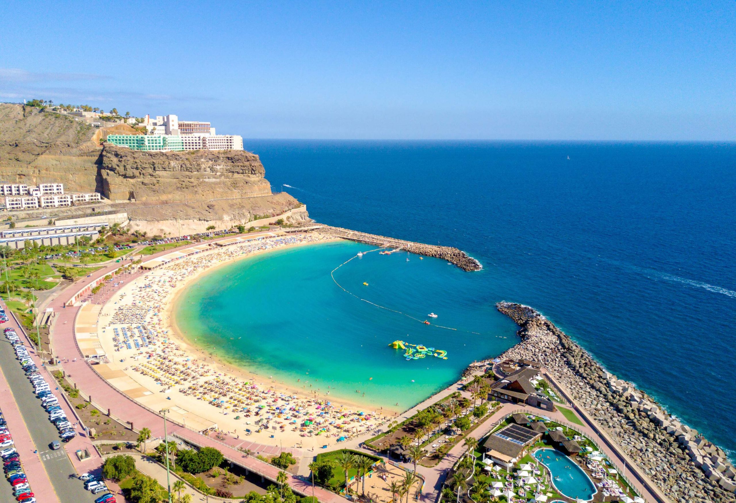 Aerial view of a crowded beach shore and the blue water beach flanked with luxury resorts and a modern building on the cliff.