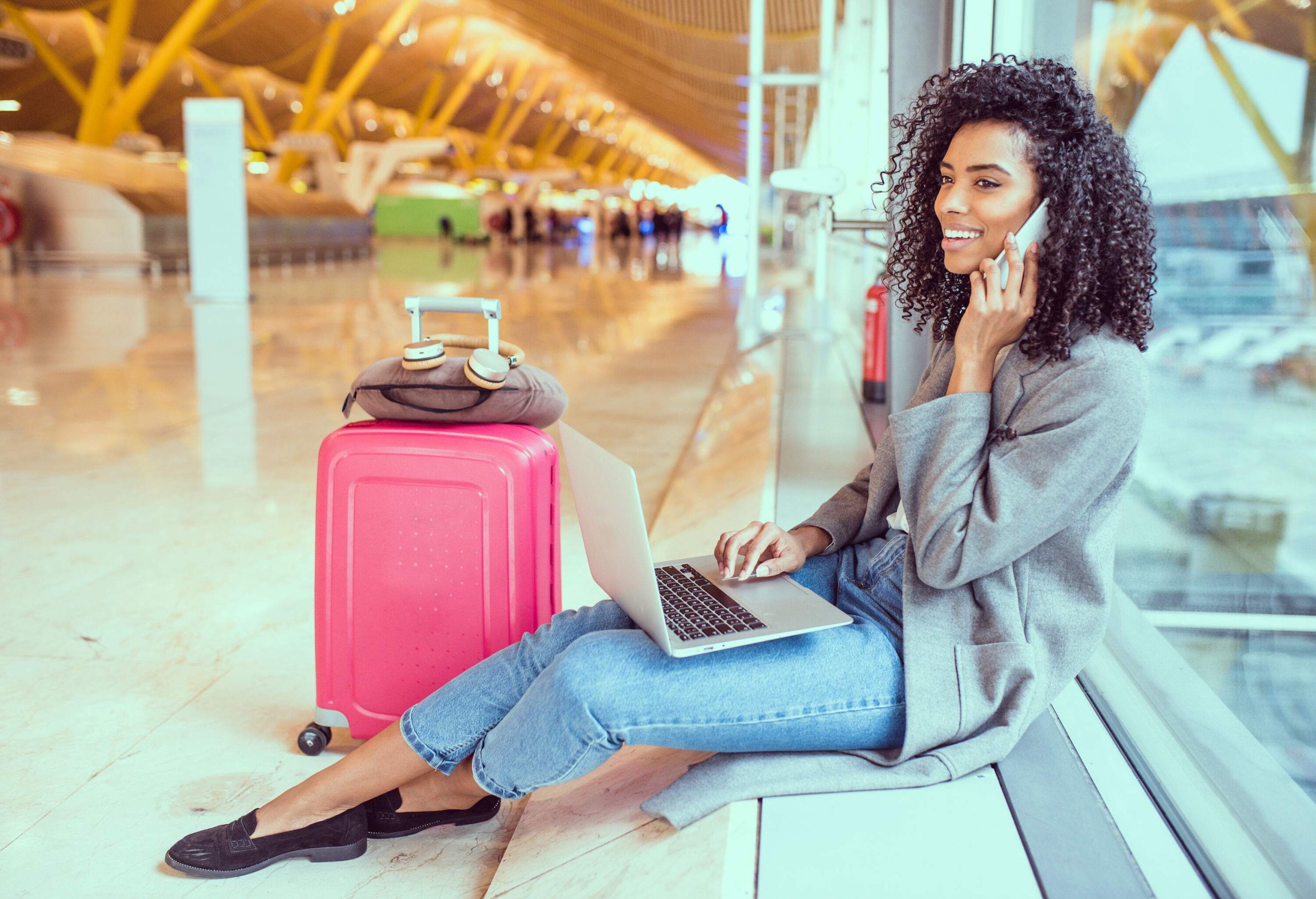 A woman in a smart casual attire sitting by the window of an airport with her laptop while talking on the phone.