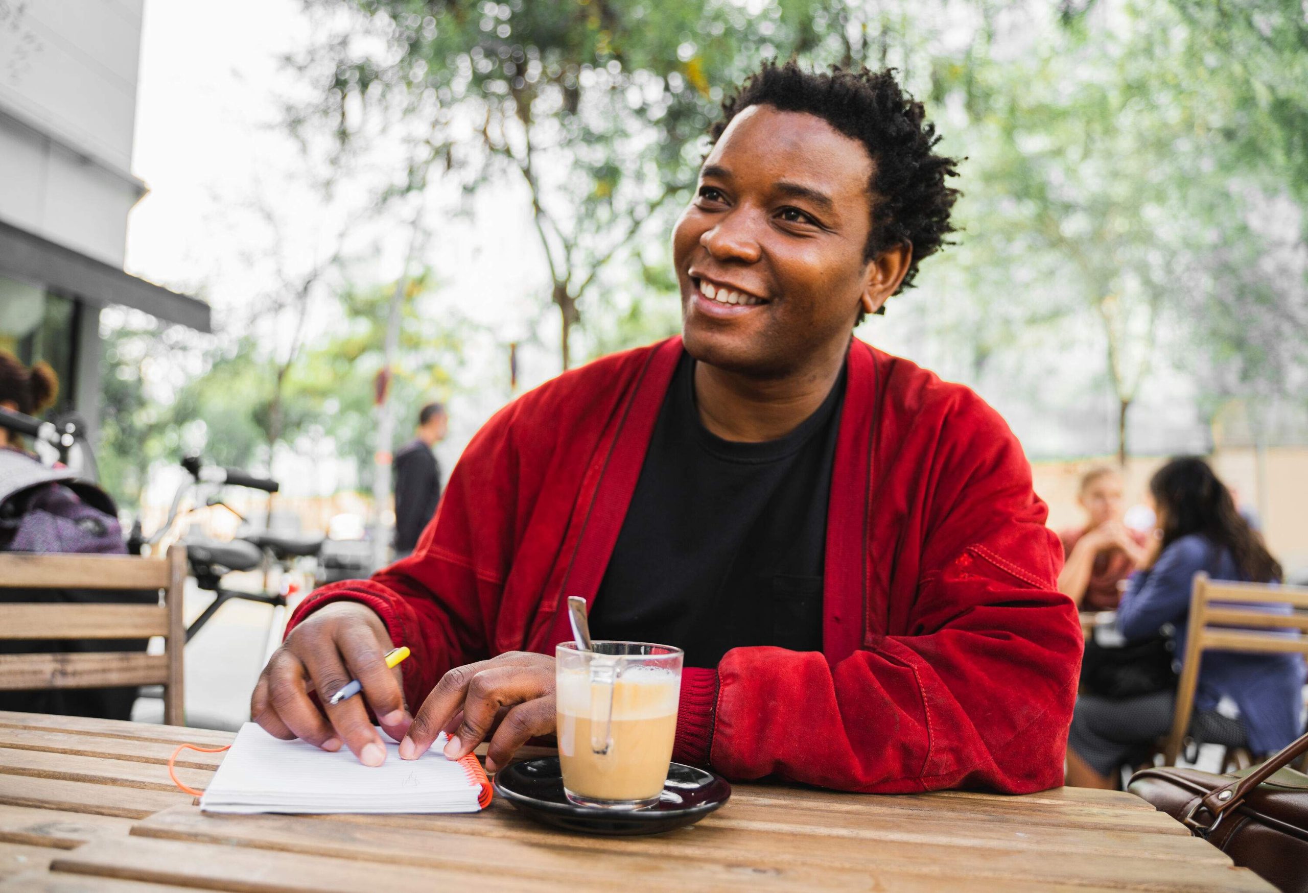 A smiling man in a red jacket holding a pen and a notebook on a table next to a cup of coffee.