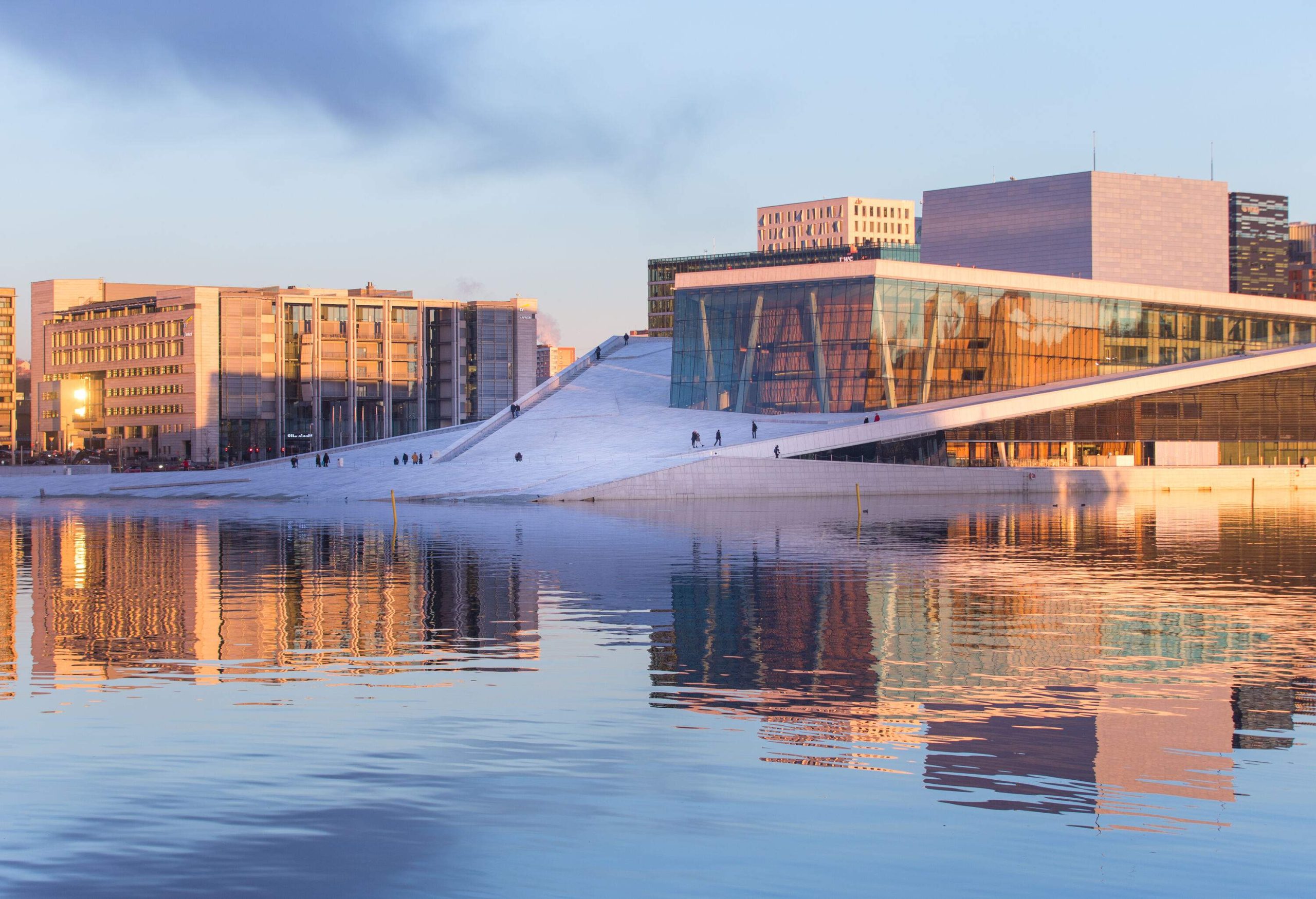 People strolling on the inclined granite surface and glass-covered Oslo Opera House façade in a waterfront neighbourhood.