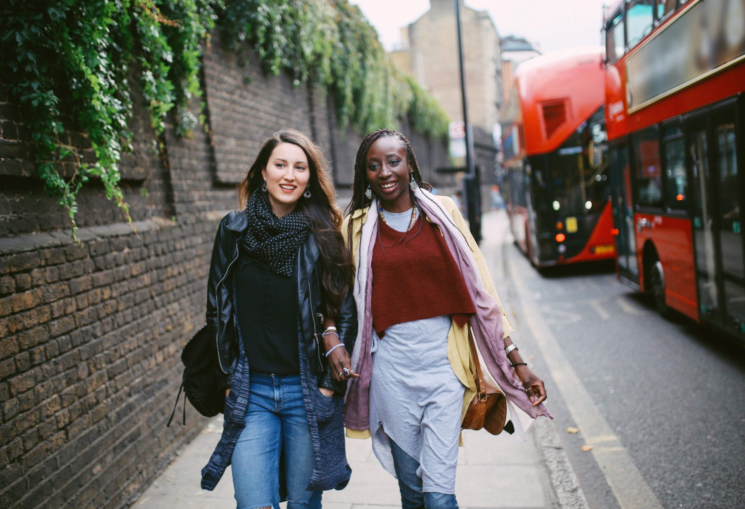 Two women in casual wear walk on the side road next to a red double-decker bus.