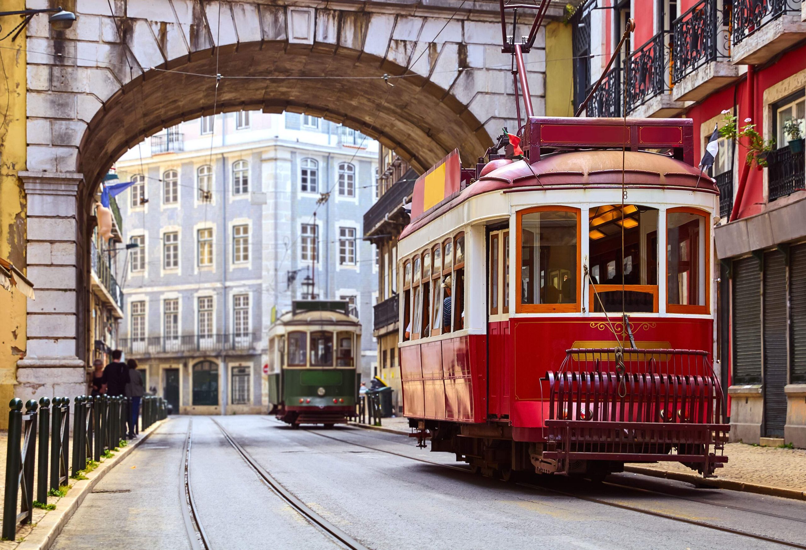 Two red vintage trams pass through an arched entrance across a street lined with old city buildings.