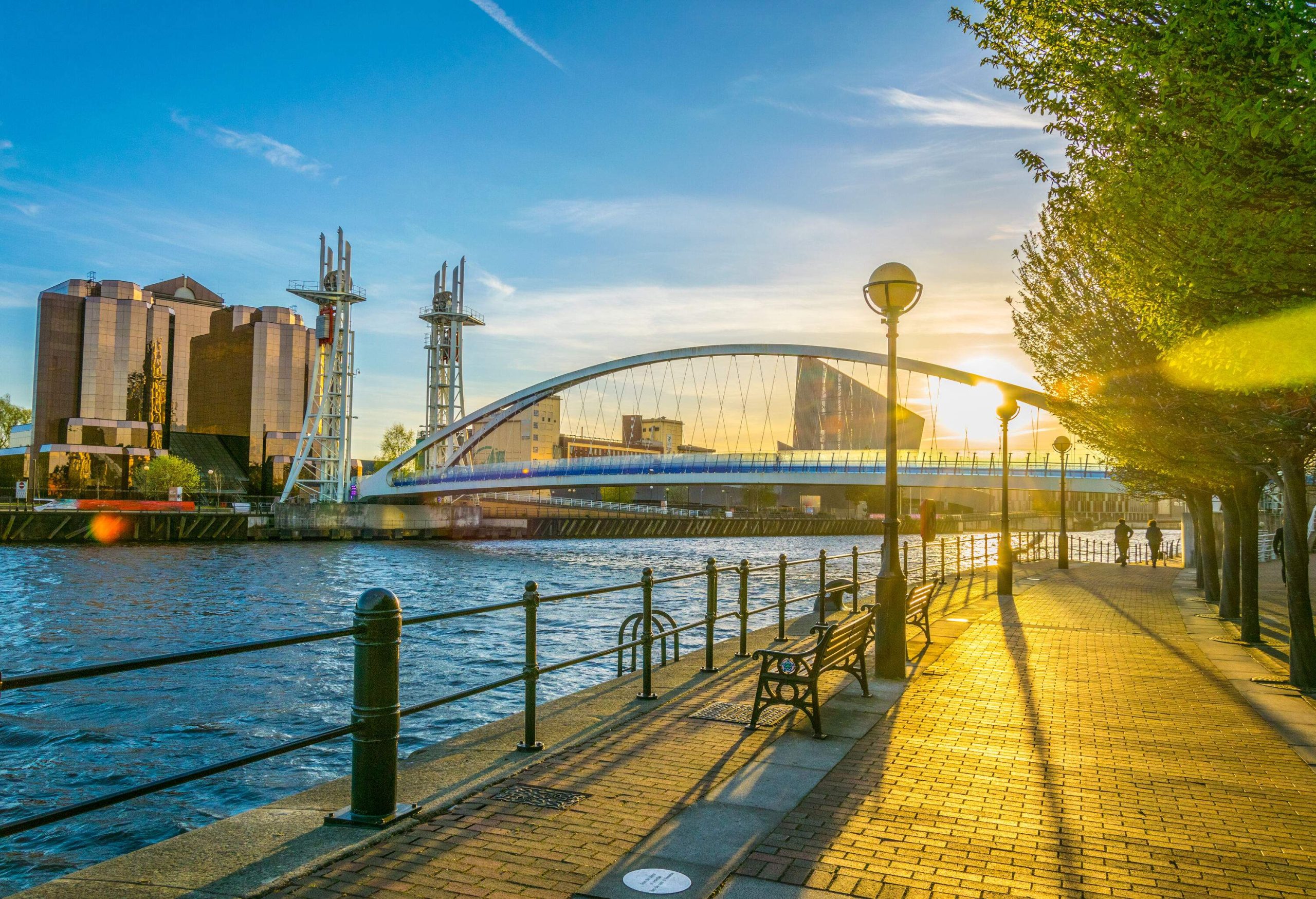 A footbridge with striking modern architecture, featuring a curved steel frame and suspension cables, spans the water in Salford Quays, Manchester, surrounded by modern buildings and a blue sky, with a promenade and guardrail in the foreground.