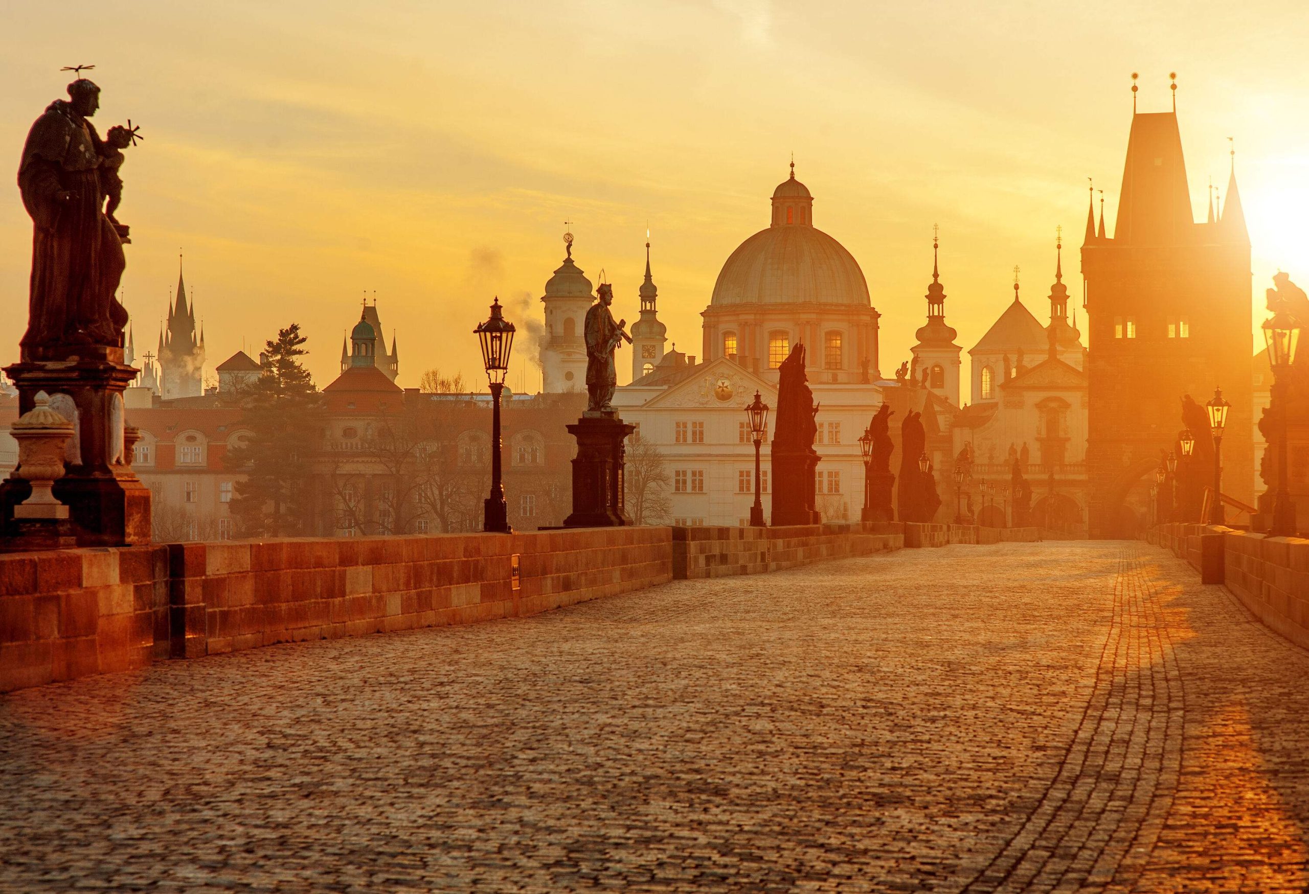 An empty paved bridge lined with statues towards the silhouette of buildings against the glaring sun.