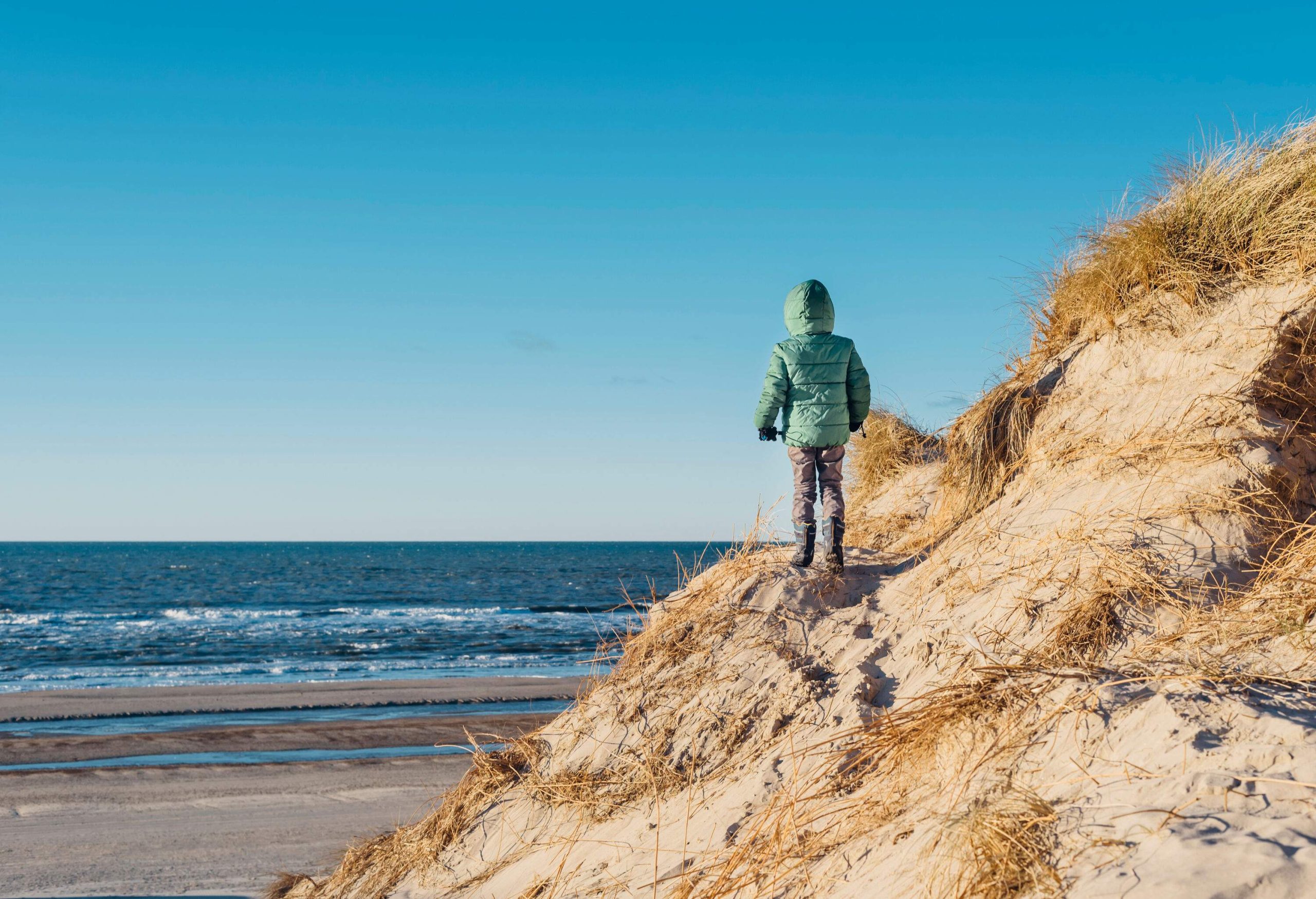 A person wearing a puffer jacket and black boots standing on a sand dune while staring at the ocean.