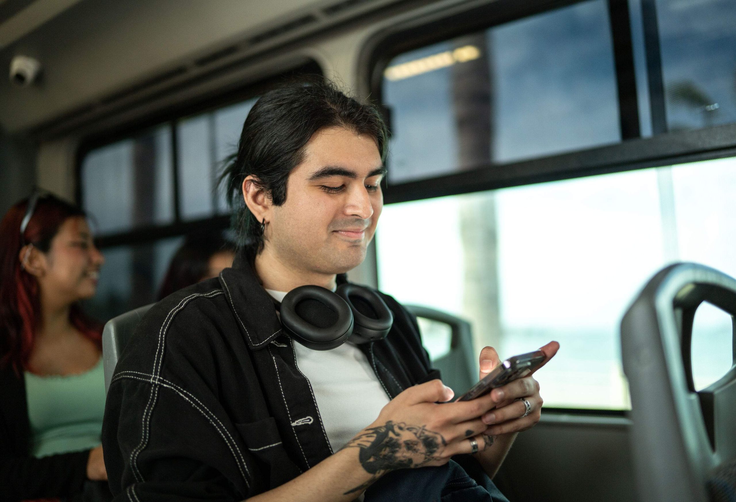 A young man on a bus wearing headphones around his neck and totally absorbed in his smartphone.