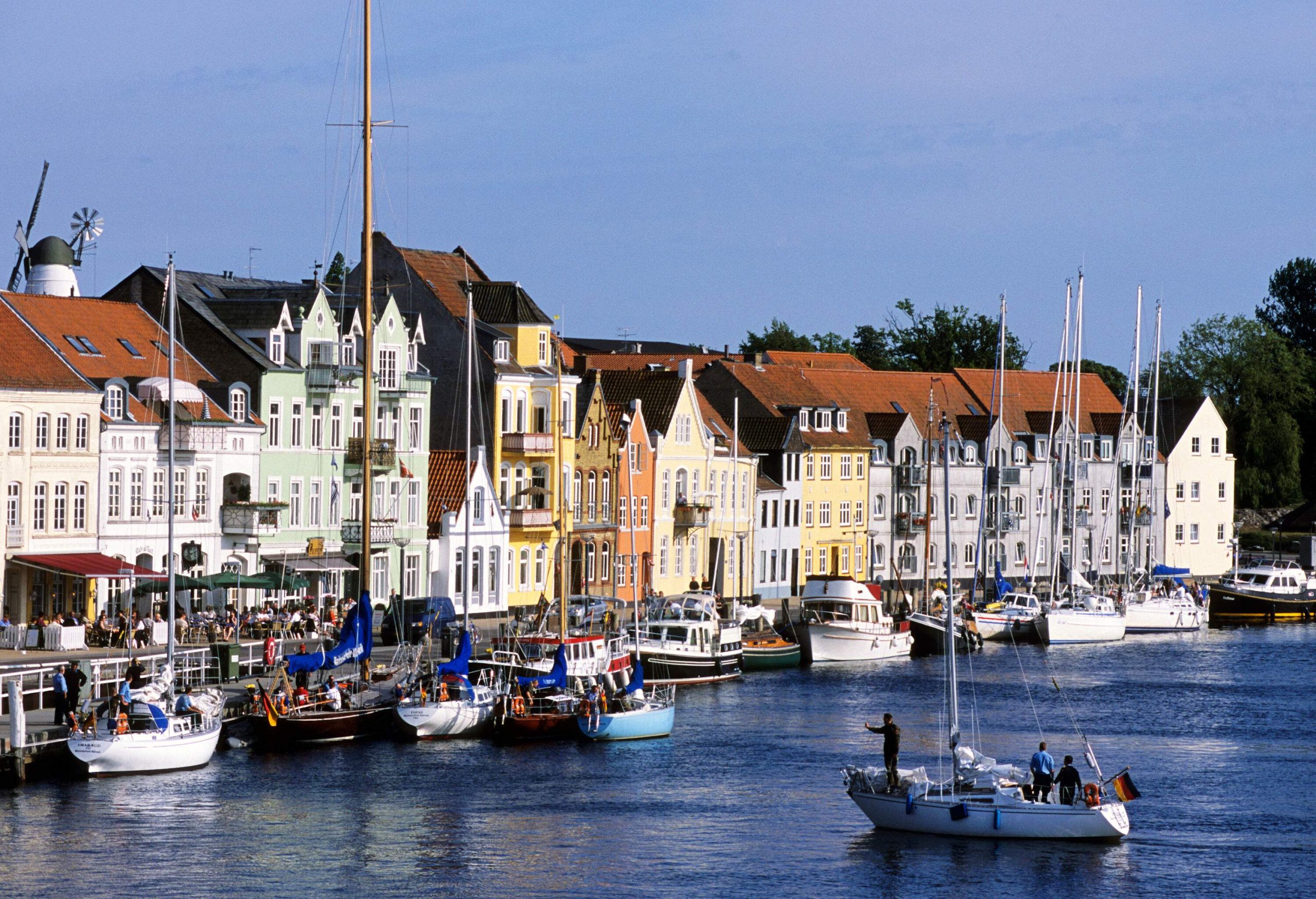 A yacht carrying passengers approaching a busy harbour backed by colourful buildings.