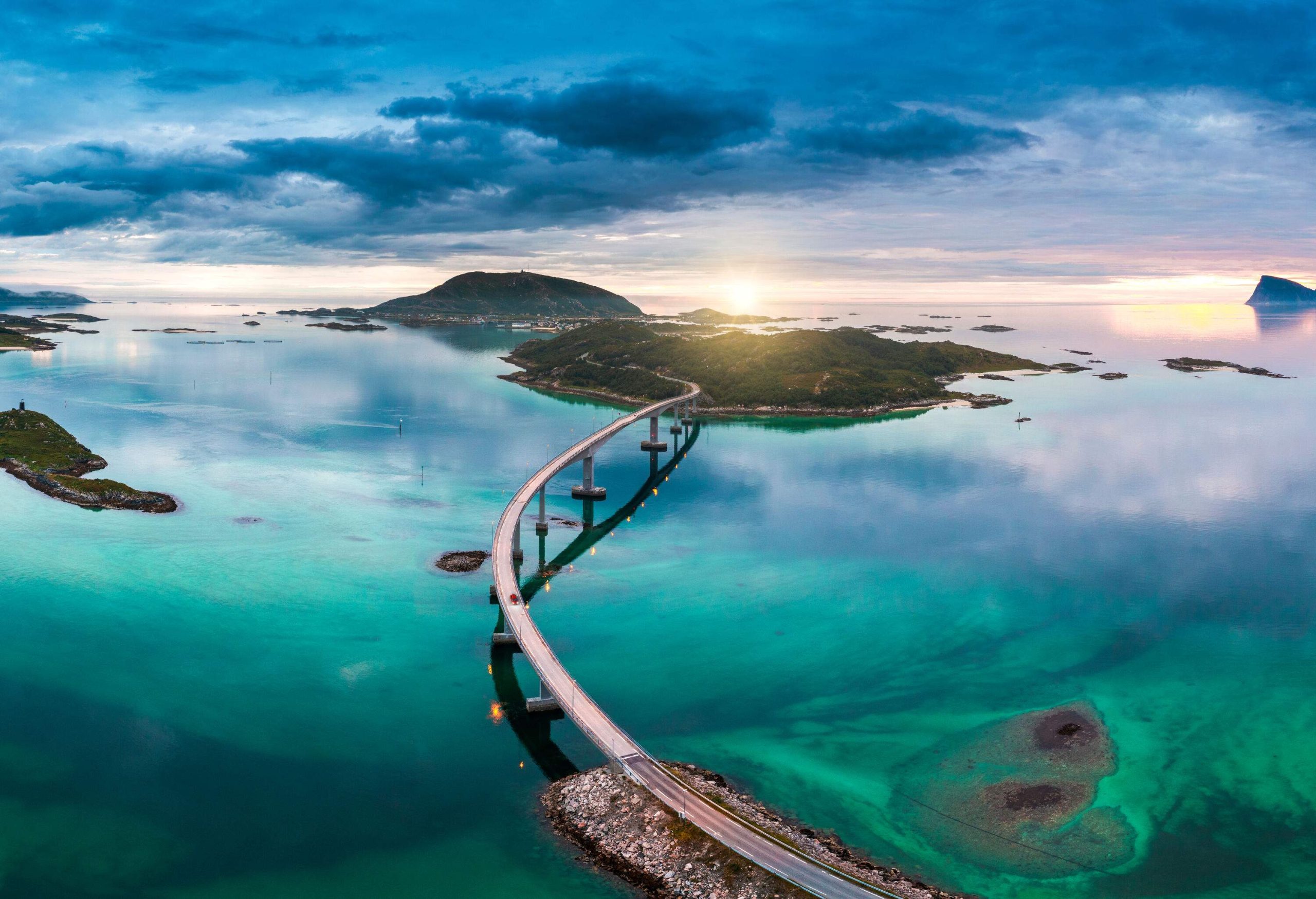 The long Tromso bridge meanders above the turquoise strait, lit by the ray of sunlight against the cloudy sky.