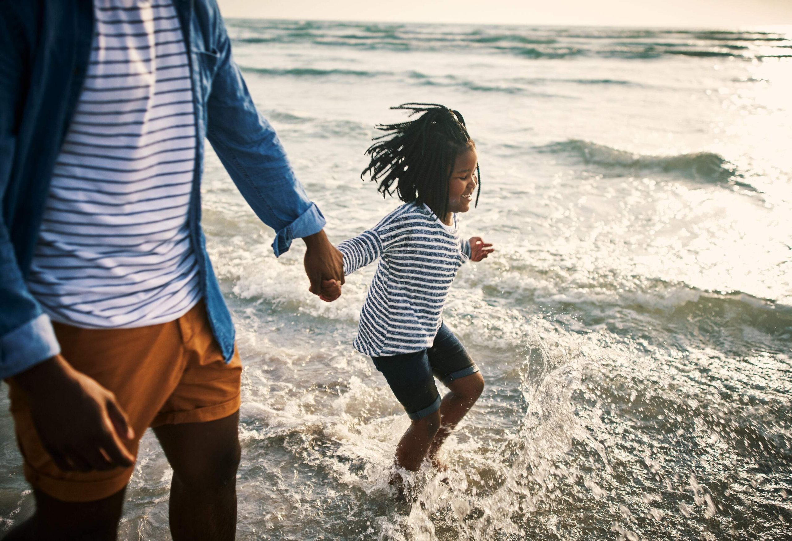 A girl in dreadlocks and striped shirt runs across the shallow waters of a beach as she hold hands with her father.