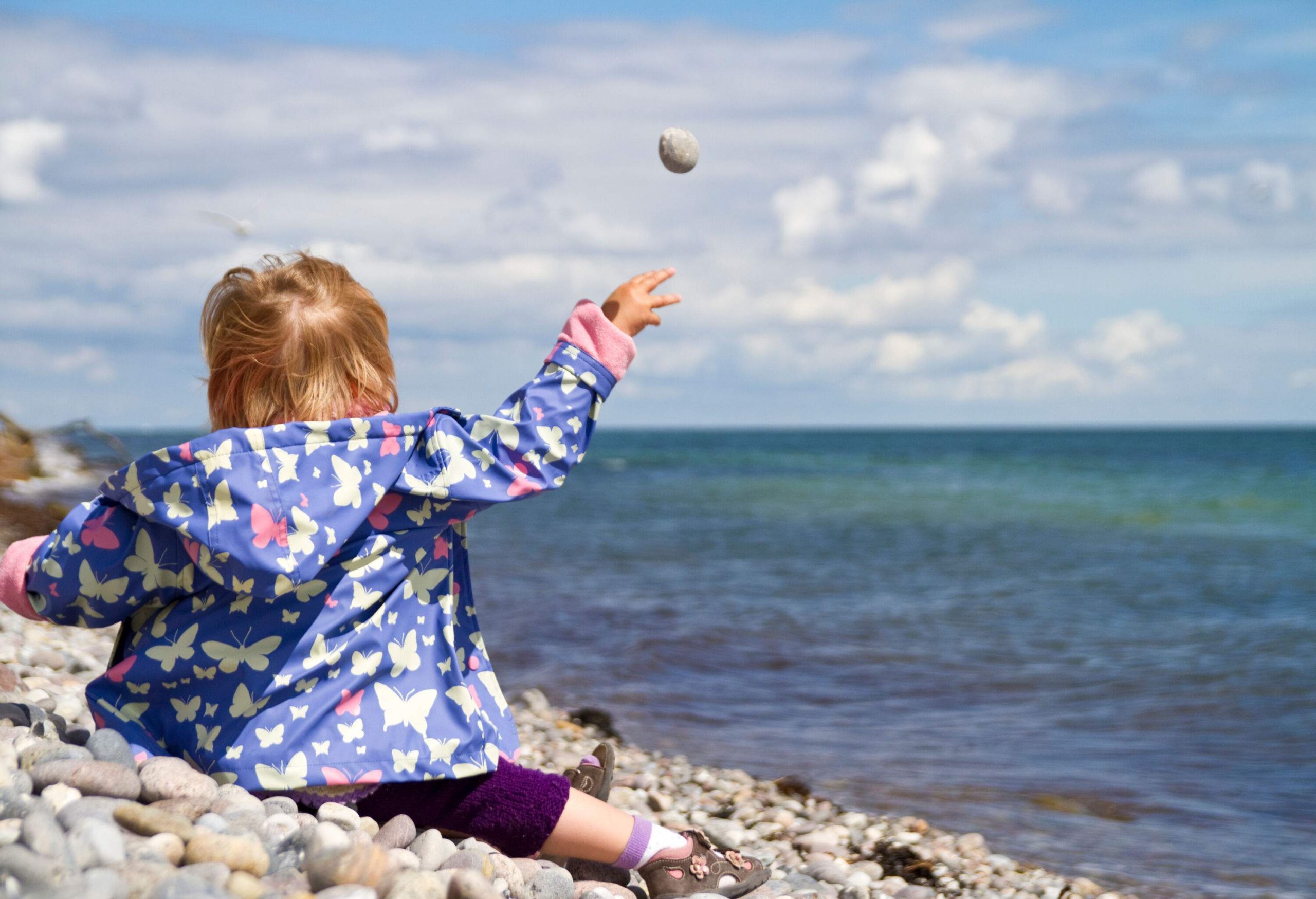 A kid in a butterfly-printed jacket sits on a pebbled beach and throws a stone.