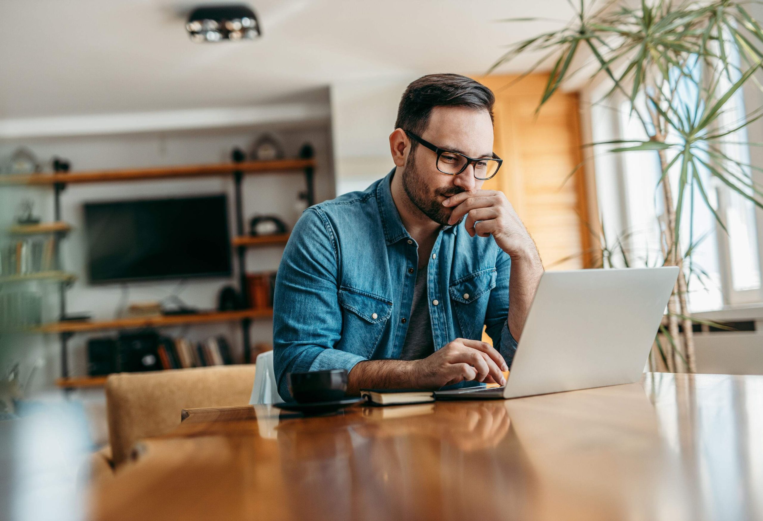 A man in eyeglasses and denim jacket sits on a table while using a laptop.