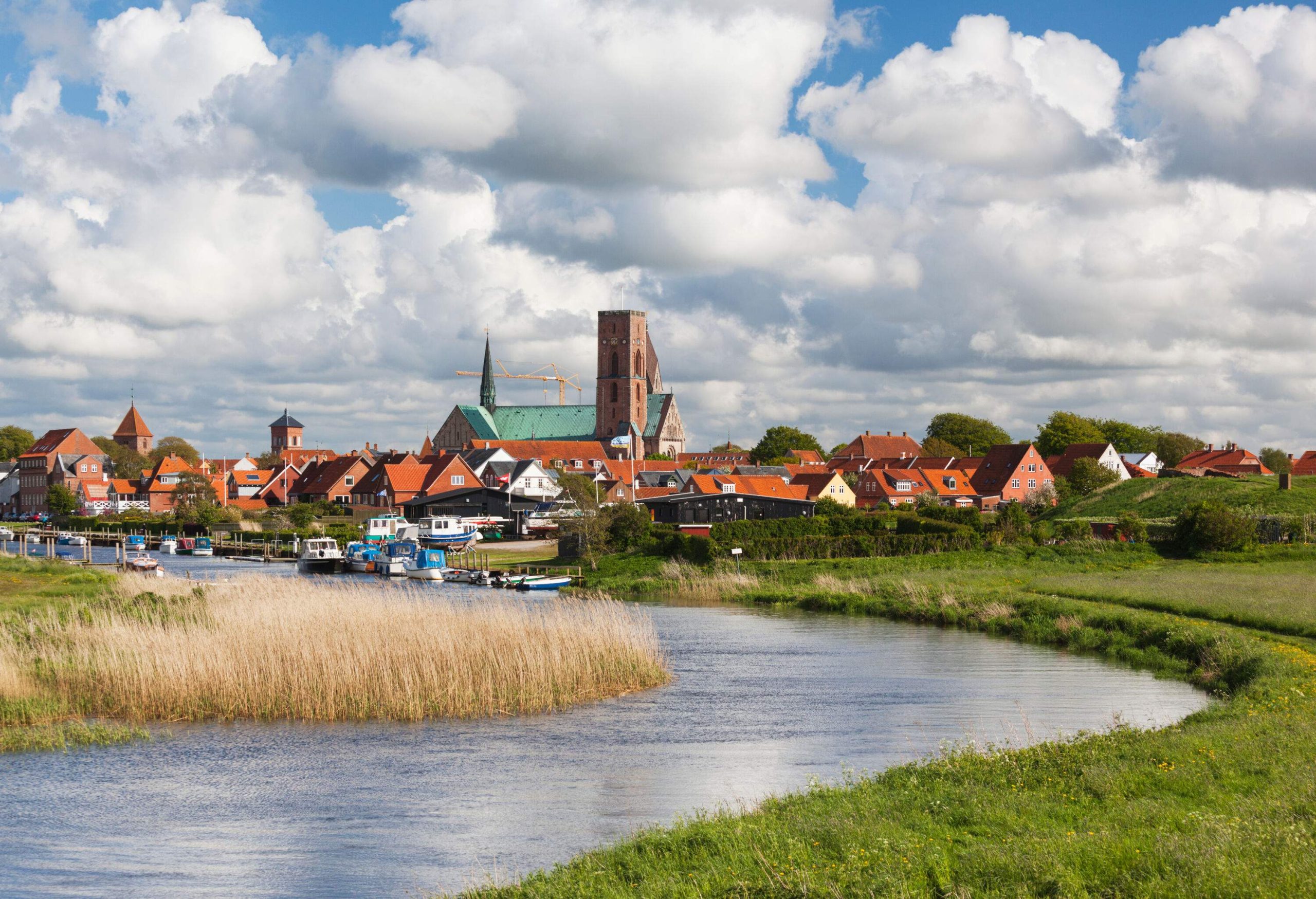 A church's square tower rises above a townscape as seen from a grassy river with anchored boats.